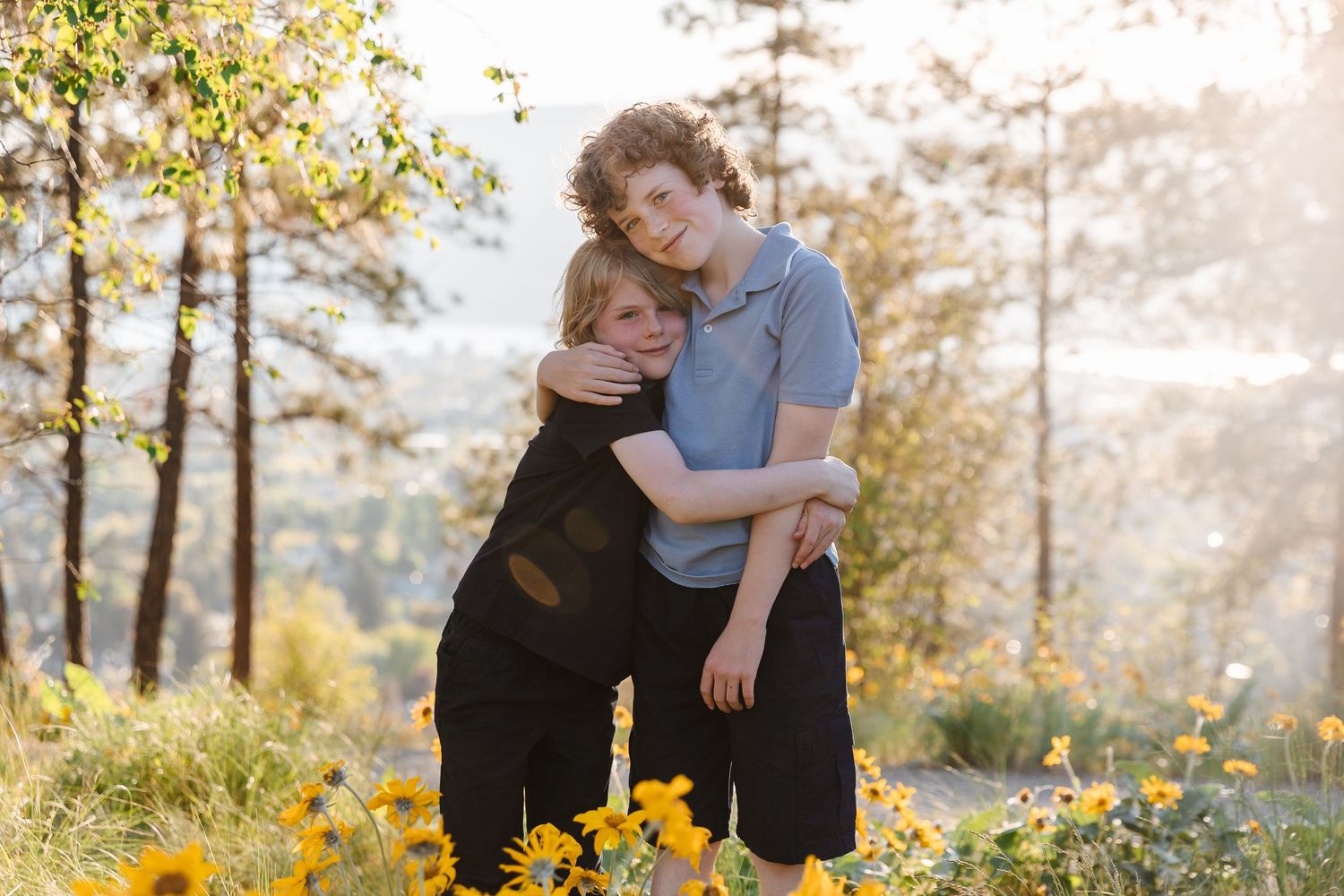 Two people share a tender embrace in a sunlit meadow with yellow wildflowers and trees in the background.