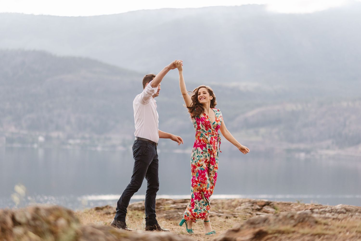 A couple dances joyfully together on a rocky shoreline with mountains and water in the background.