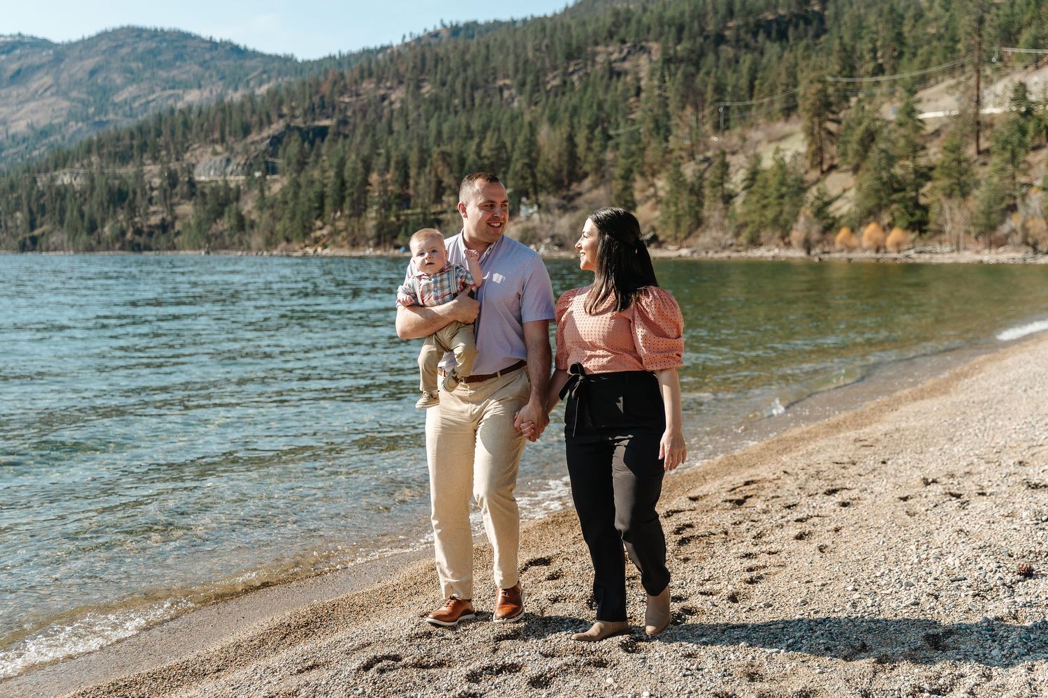 Couple walking along a scenic mountain lake shoreline with evergreen trees and rocky cliffs in the background.