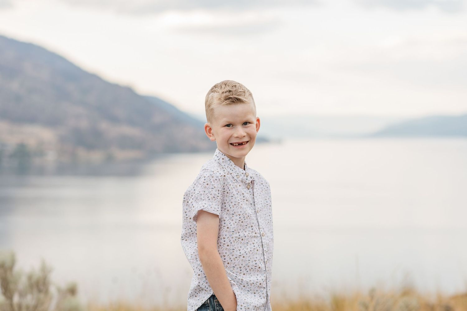 A scenic family photoshoot takes place on a grassy hillside overlooking a serene lake with mountains in the background.