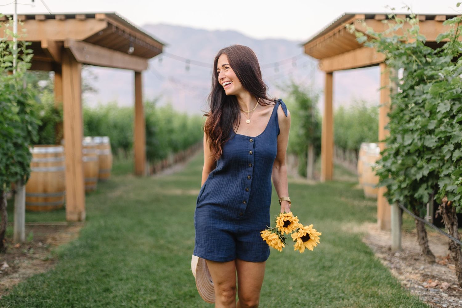 A person in a blue romper walks through a vineyard holding sunflowers with wooden pergolas and mountains in the background.