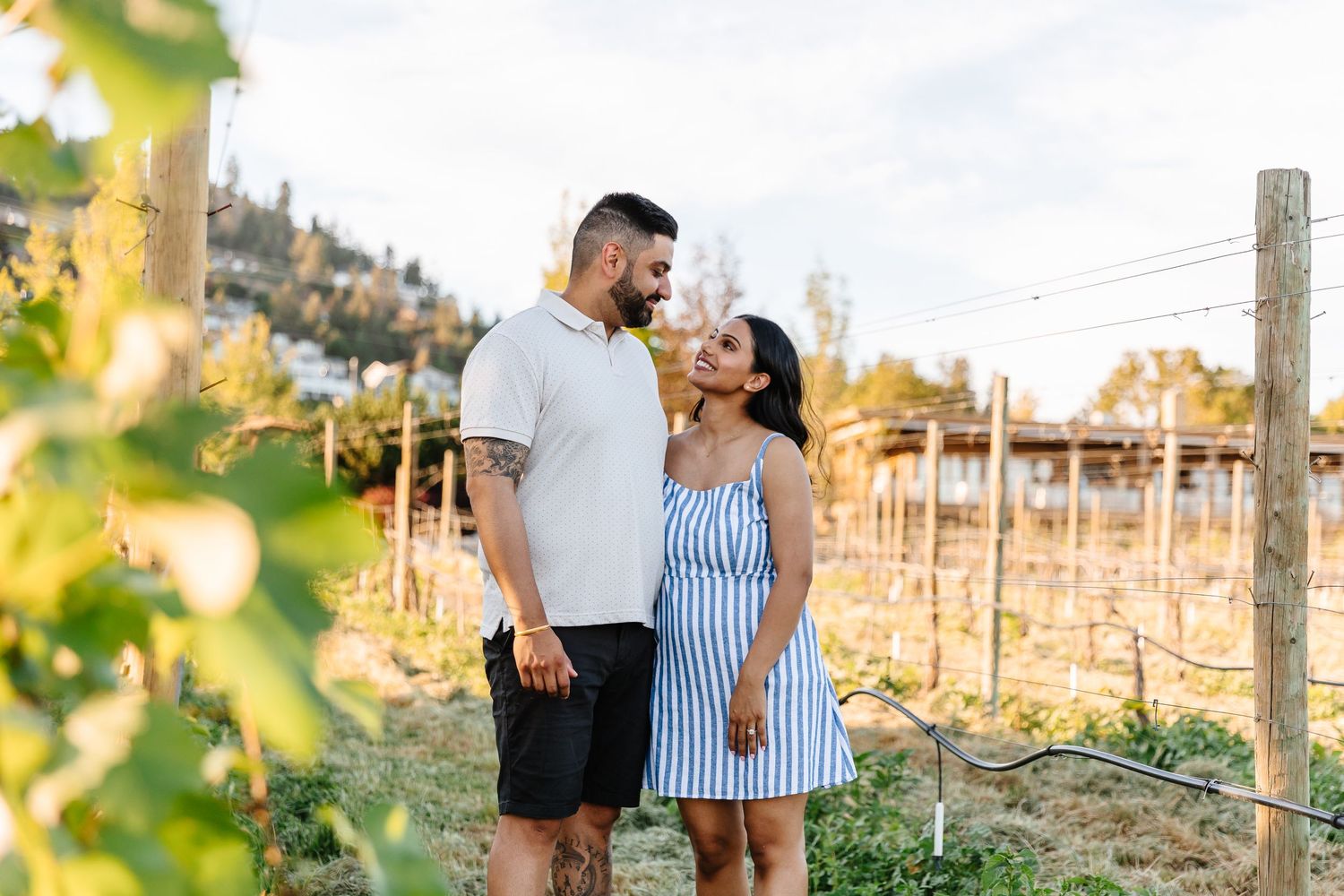 Couple walking through scenic vineyard at sunset holding wine glasses and enjoying romantic moment together.