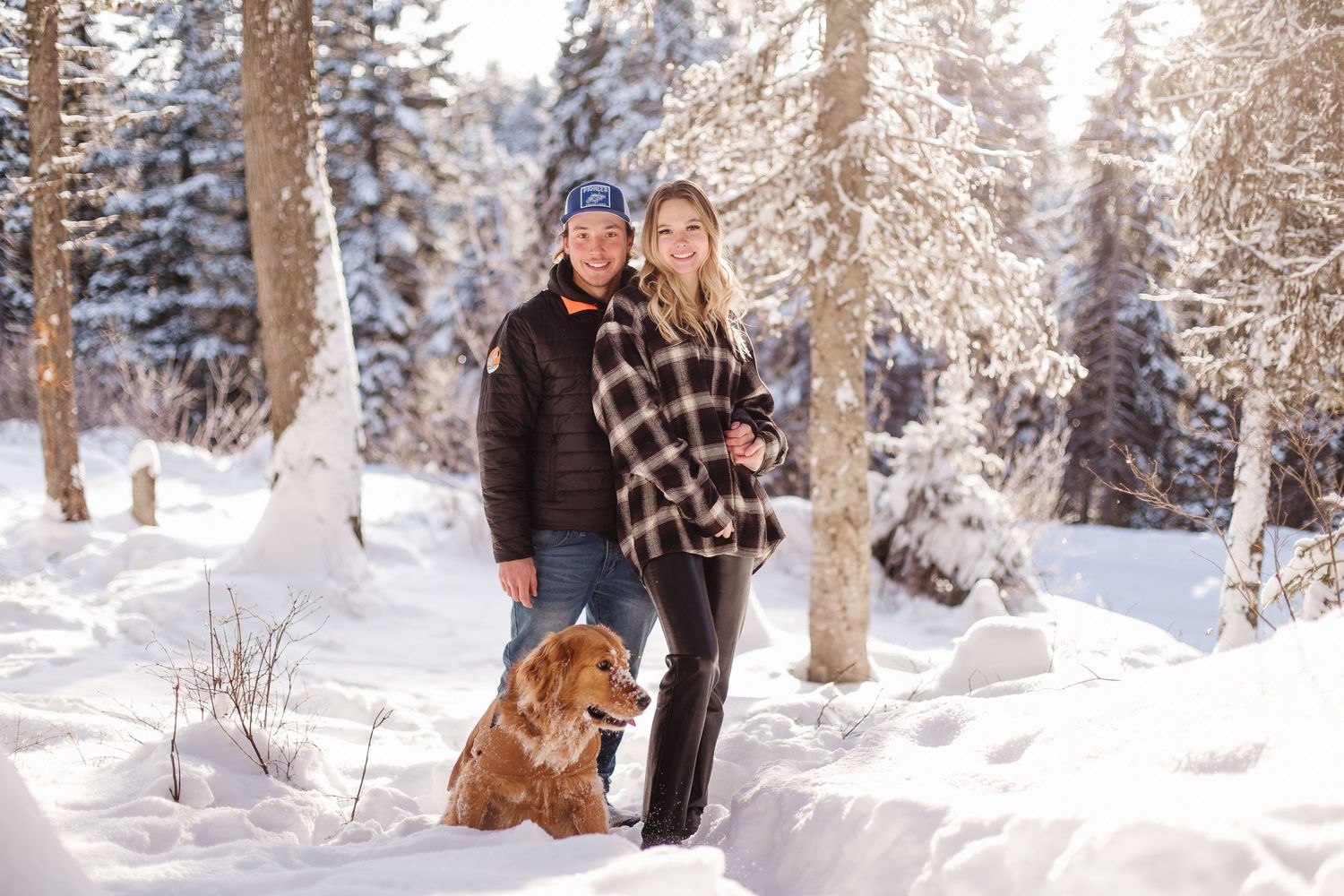 Two people and their golden retriever dog pose together in a snowy winter forest landscape on a sunny day.