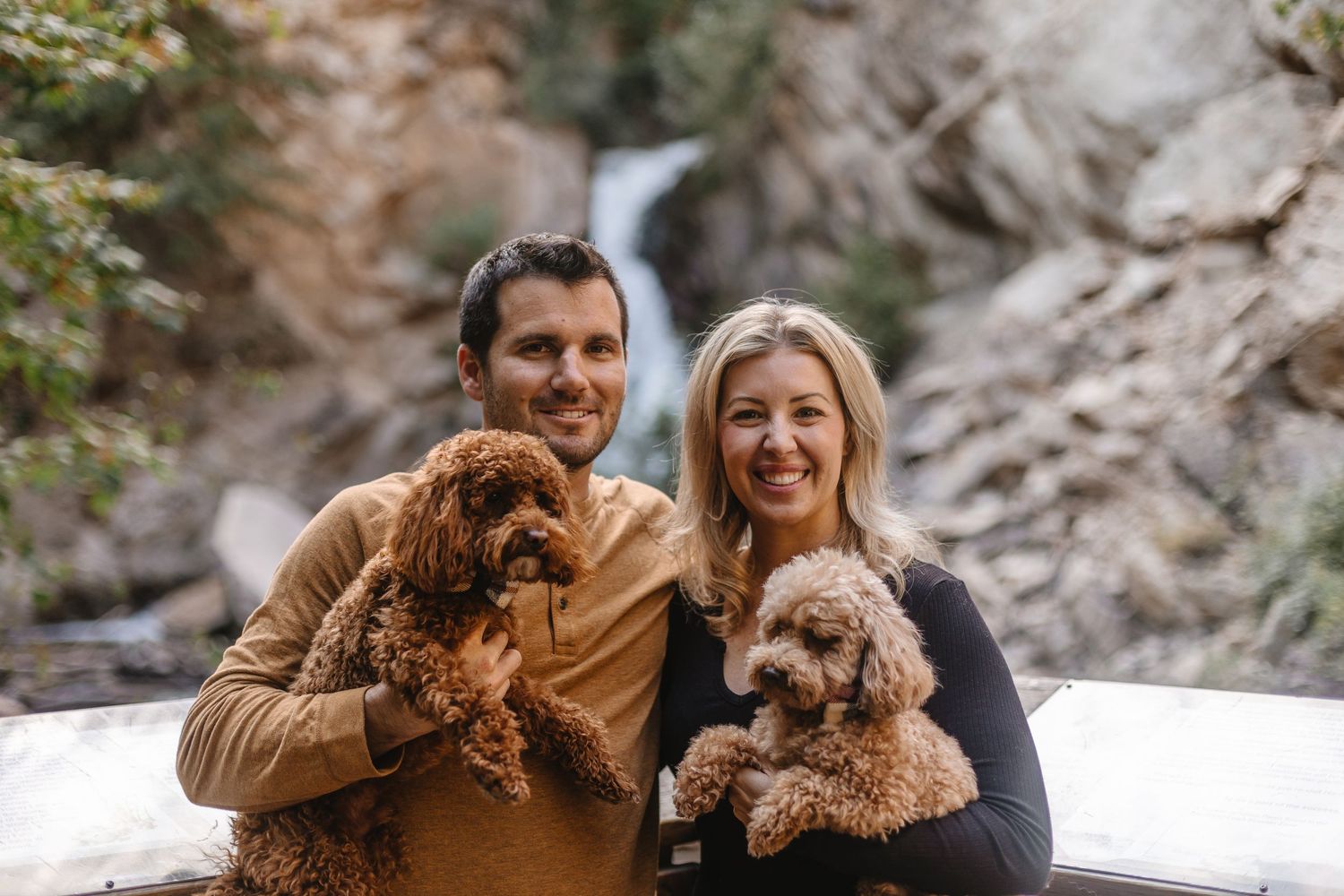 A couple holds their small brown dogs while posing in front of a scenic waterfall in the mountains.