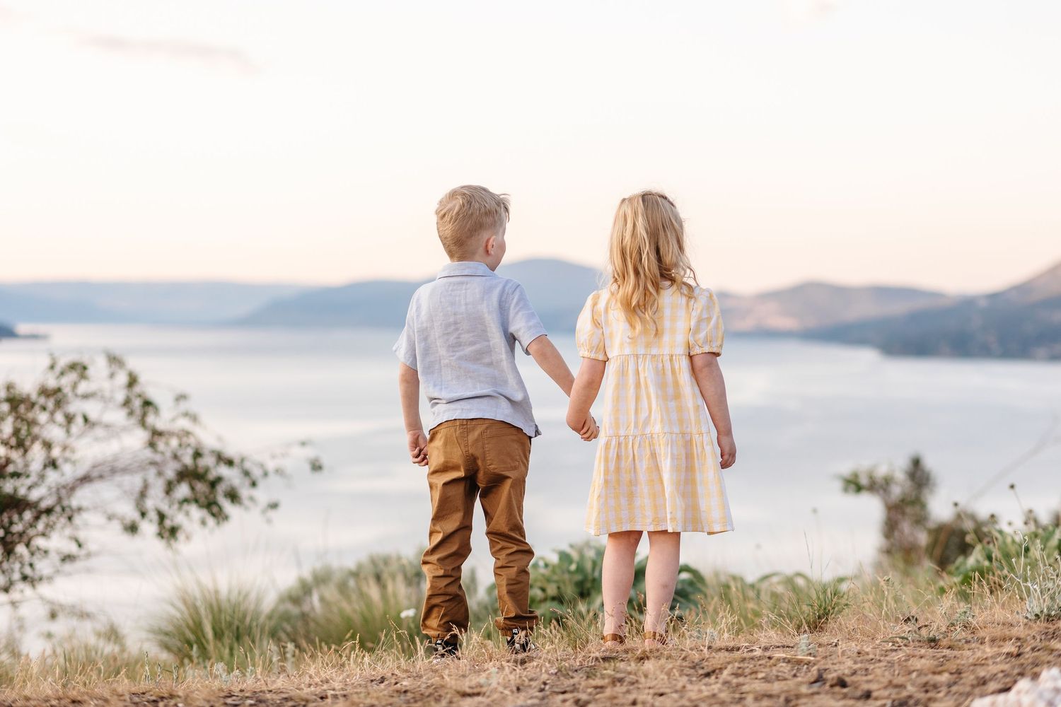 Two children hold hands while standing on a hilltop overlooking a misty lake and mountains in the distance.