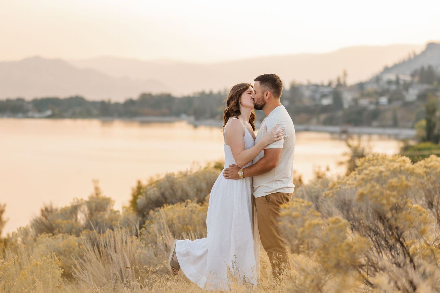 A couple embraces tenderly during a golden hour photoshoot in a scenic outdoor setting with tall grass and warm sunlight.