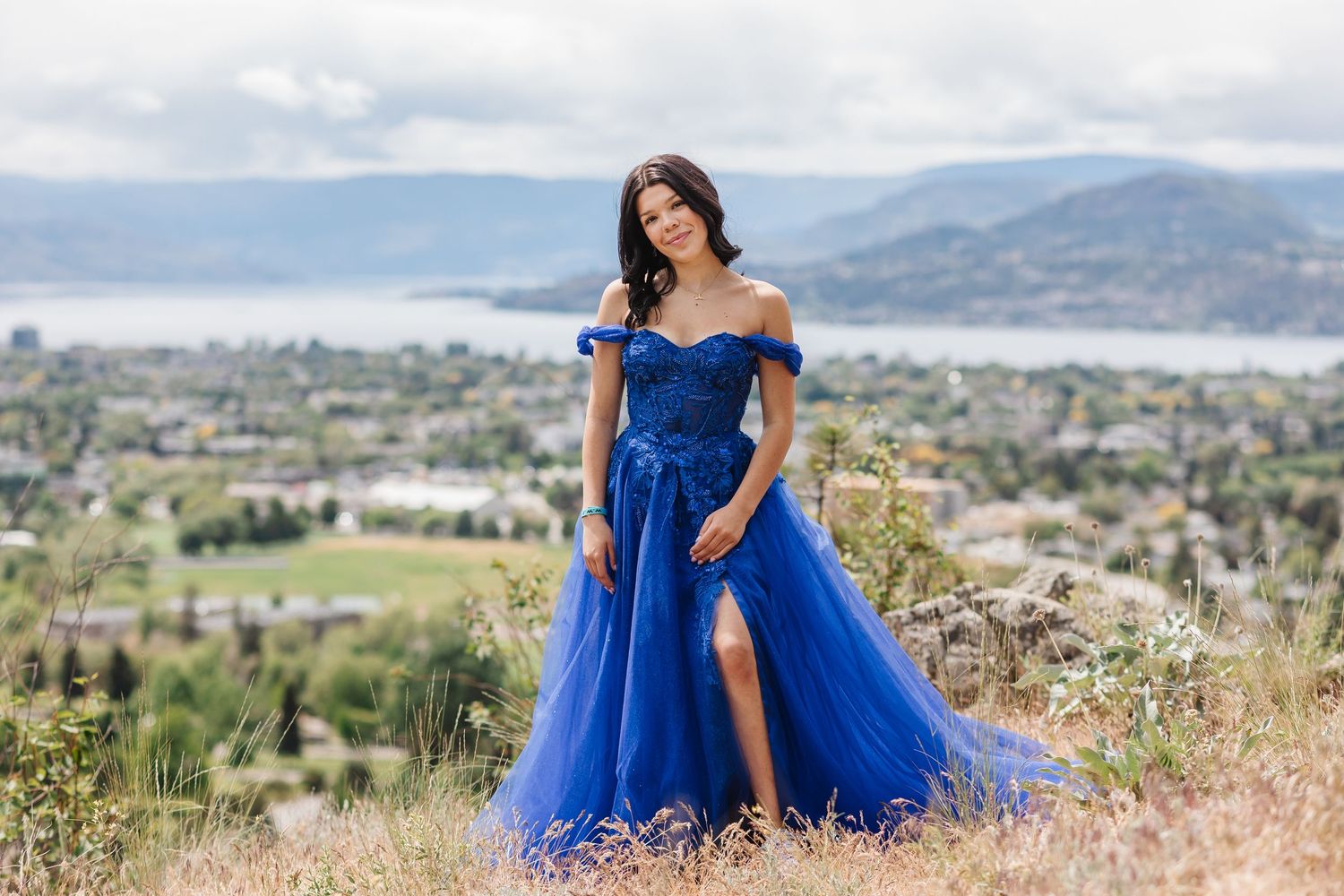 A person in a flowing royal blue gown poses gracefully in a scenic mountain landscape with lake views.