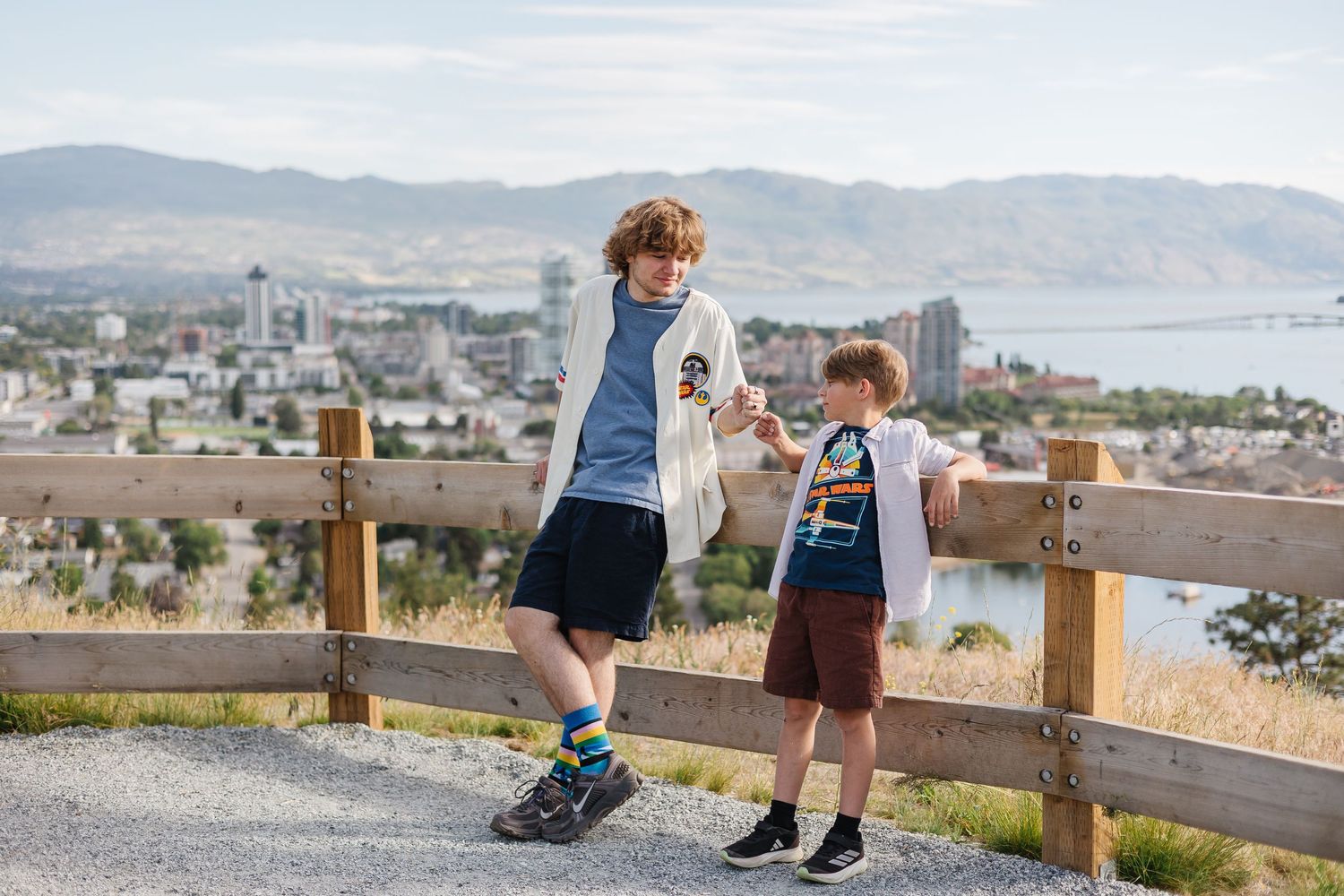 Children's feet dangle from wooden bench overlooking scenic mountain vista and city landscape below.