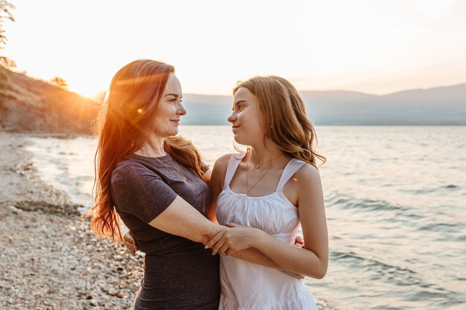 A couple embraces lovingly on a beach at sunset with mountains and ocean in the background.
