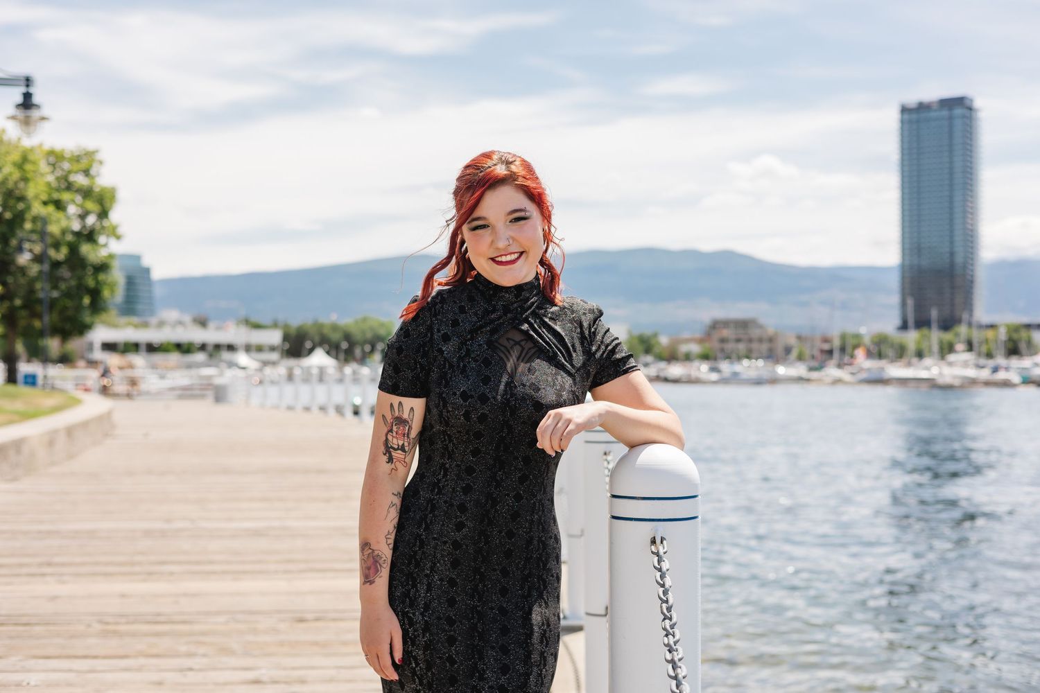 Woman in black dress walking along waterfront path with mountains and tall buildings in background.