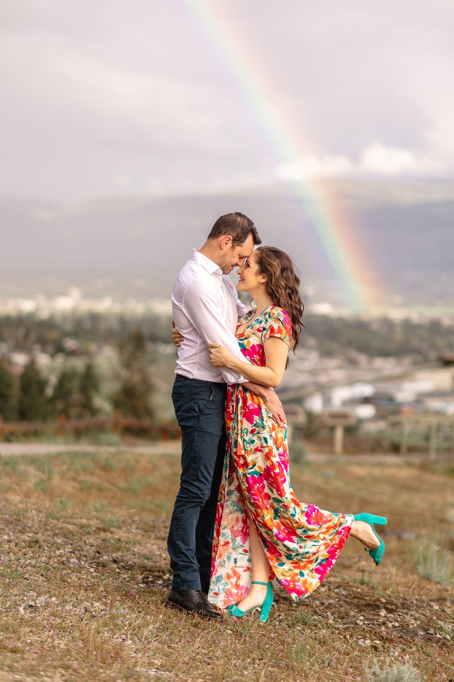 A couple embraces romantically in a field with a vibrant rainbow arching across the cloudy sky in the background.
