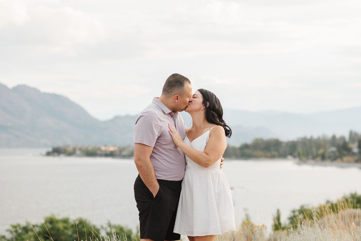 A couple shares a romantic kiss against a scenic mountain lake backdrop during their outdoor photo session.