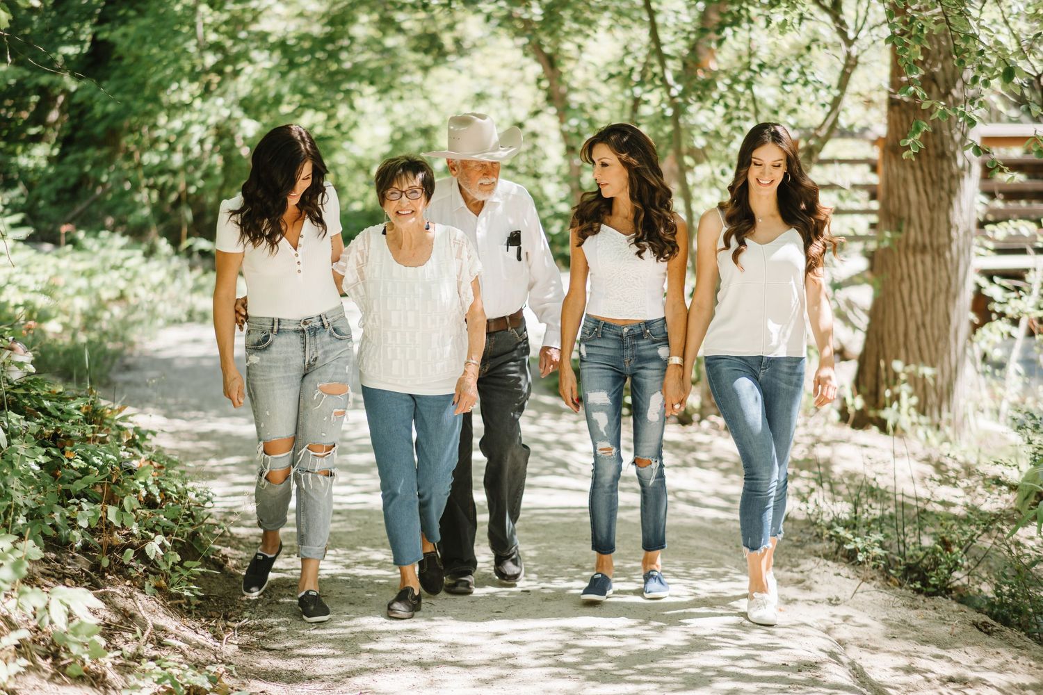 A group of people in casual denim jeans and white tops walking together on a tree-lined path in summer sunlight.