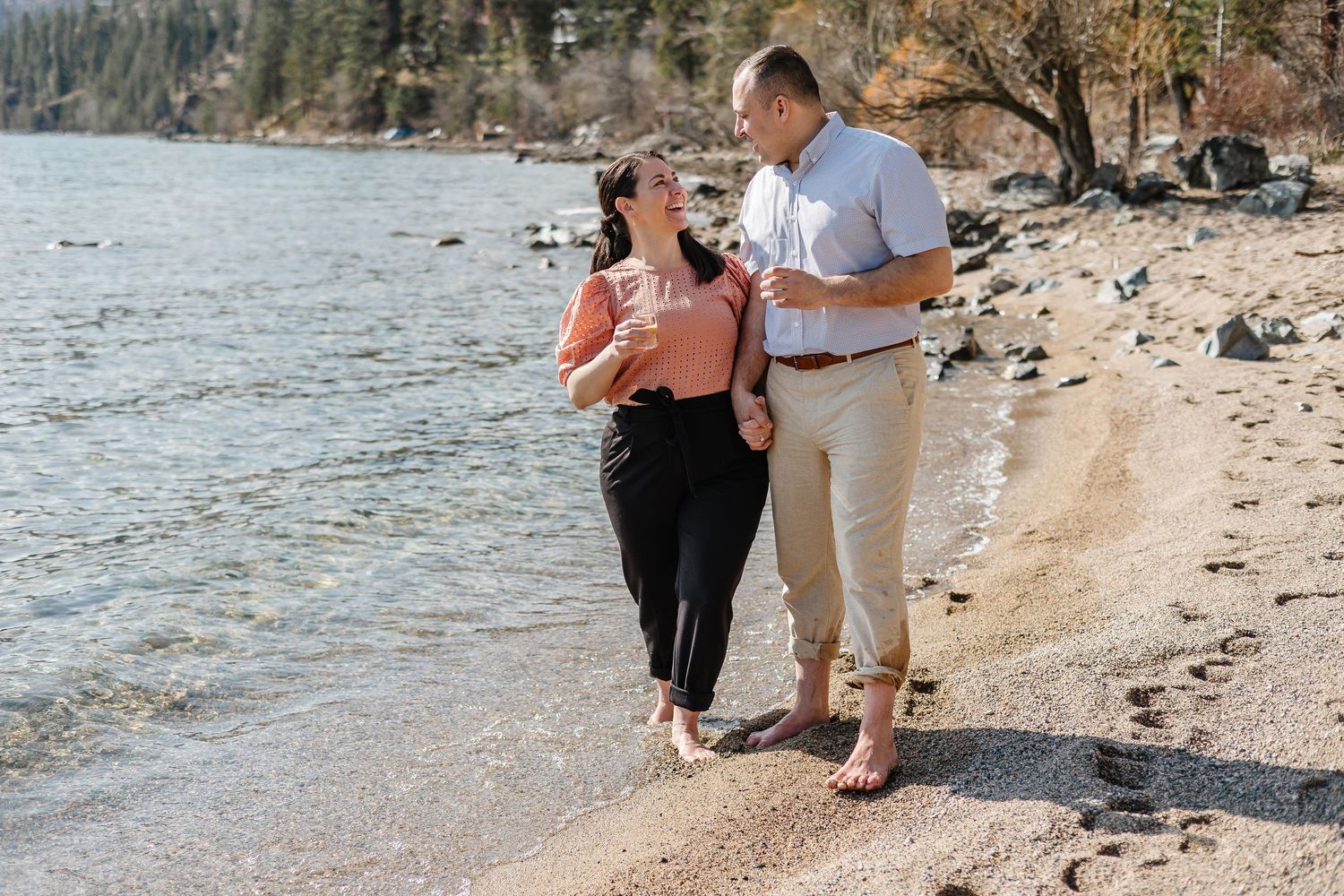 Couple walking barefoot along a rocky beach shoreline during a sunny day.