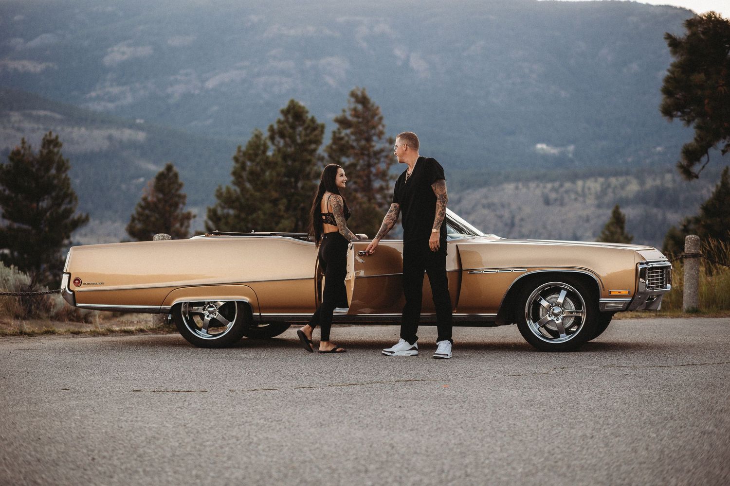 A vintage gold Cadillac convertible parked on a scenic mountain overlook with mountains in the distance.