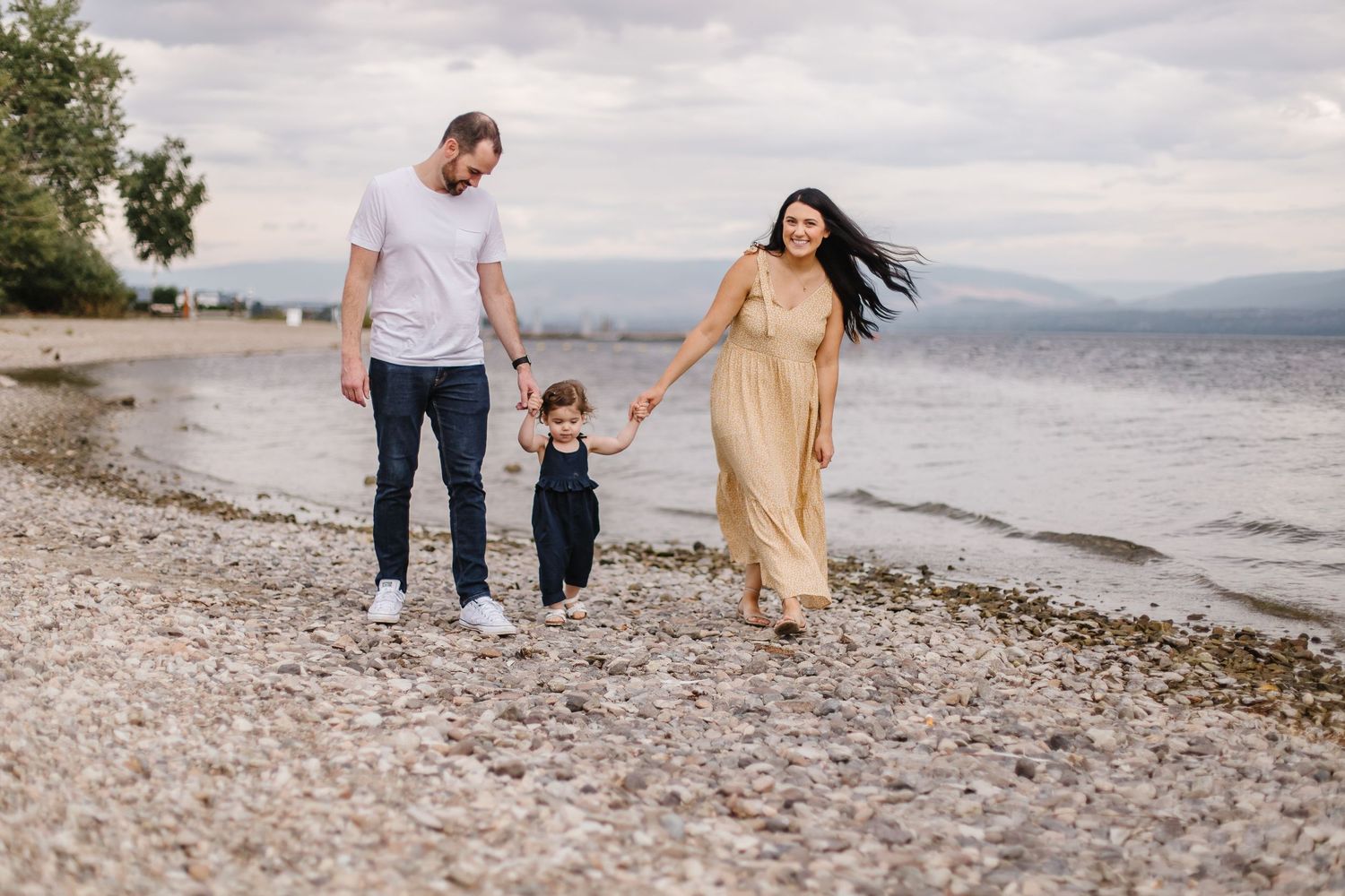A couple and child walk along a rocky beach shoreline during an overcast day with mountains in the distance.