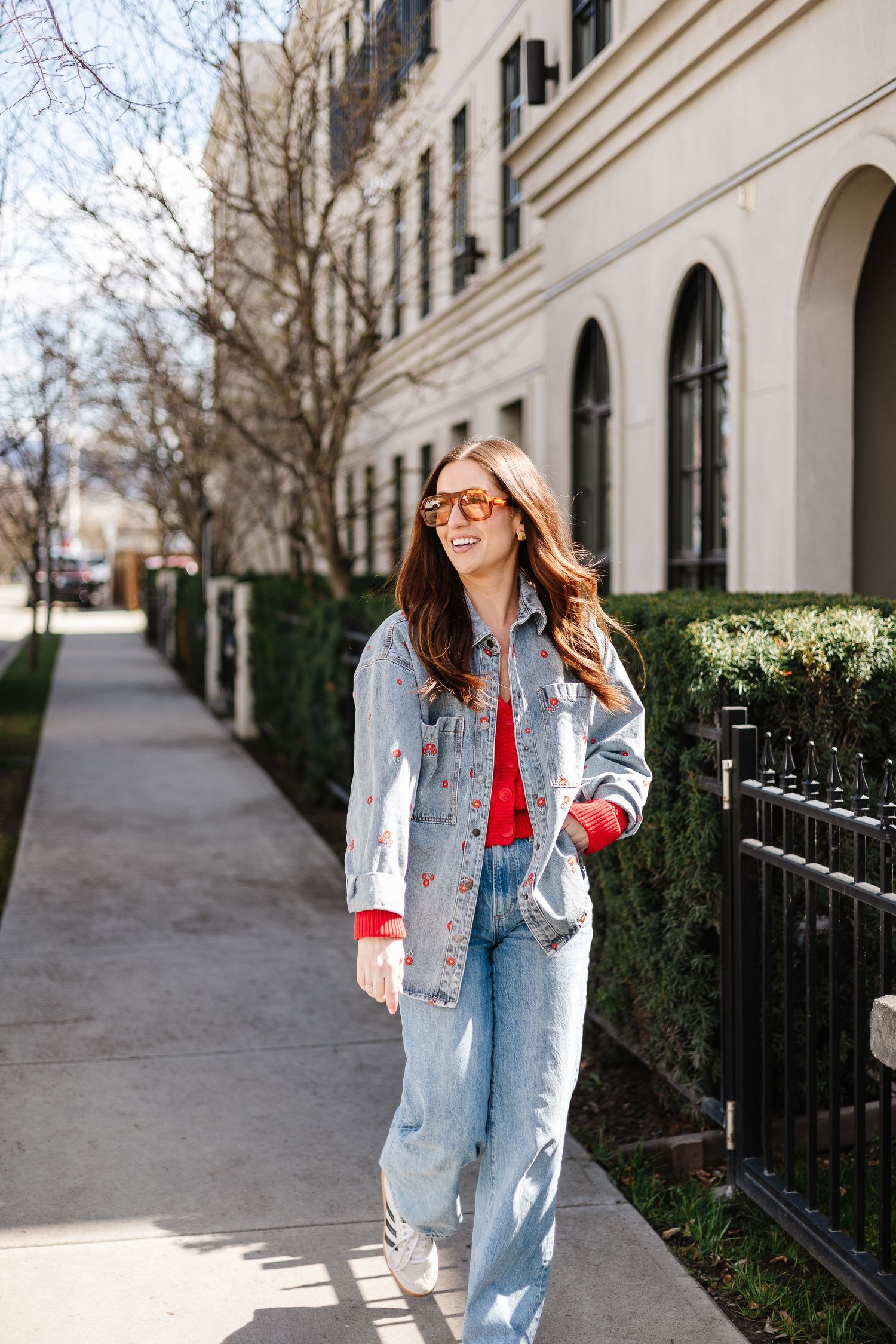 Stylish person wearing light denim jacket and red top poses on city sidewalk next to black iron fence and buildings.