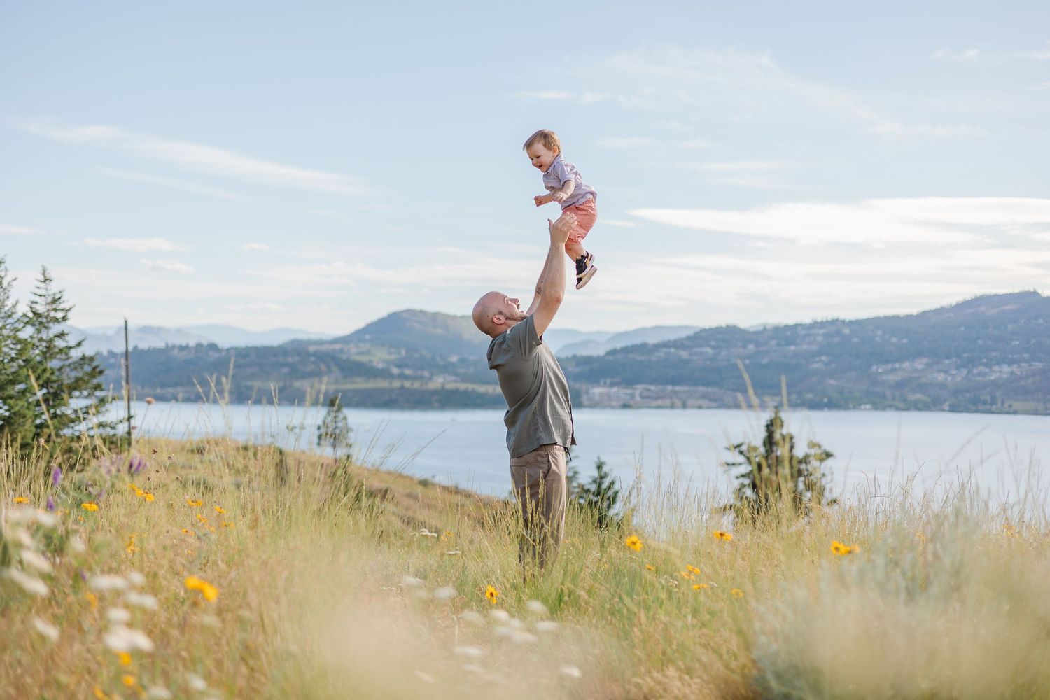 Person stands on grassy hillside overlooking scenic lake and mountain landscape on sunny day.