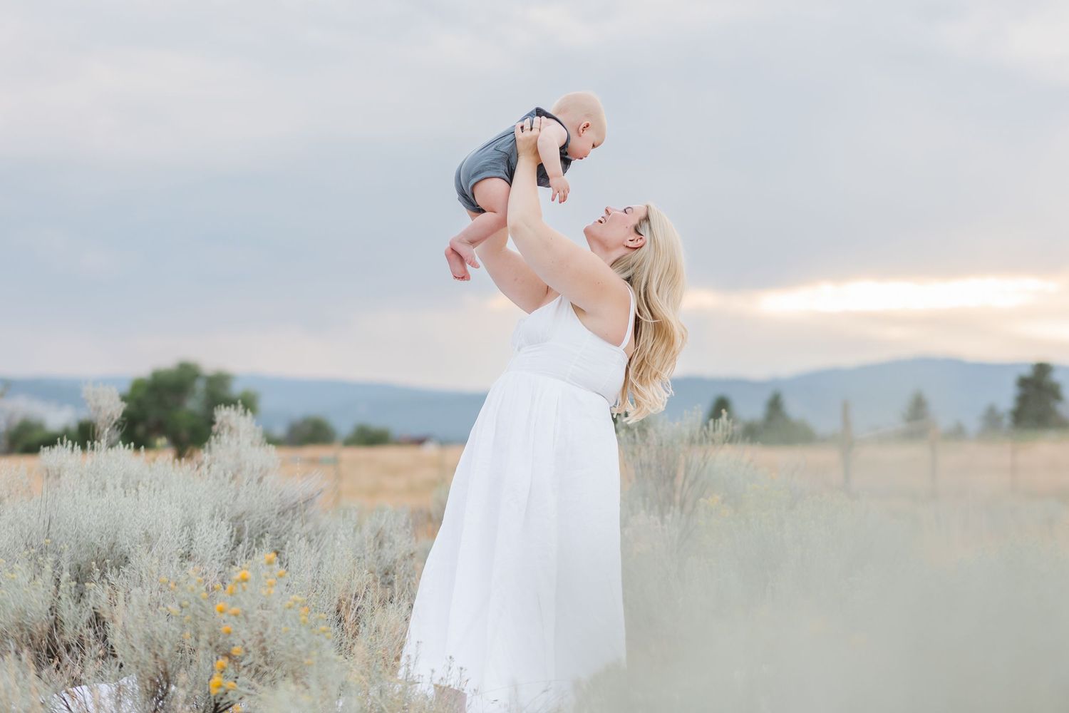 Parent in flowing white dress lifts baby joyfully in desert field during sunset with mountains in background.