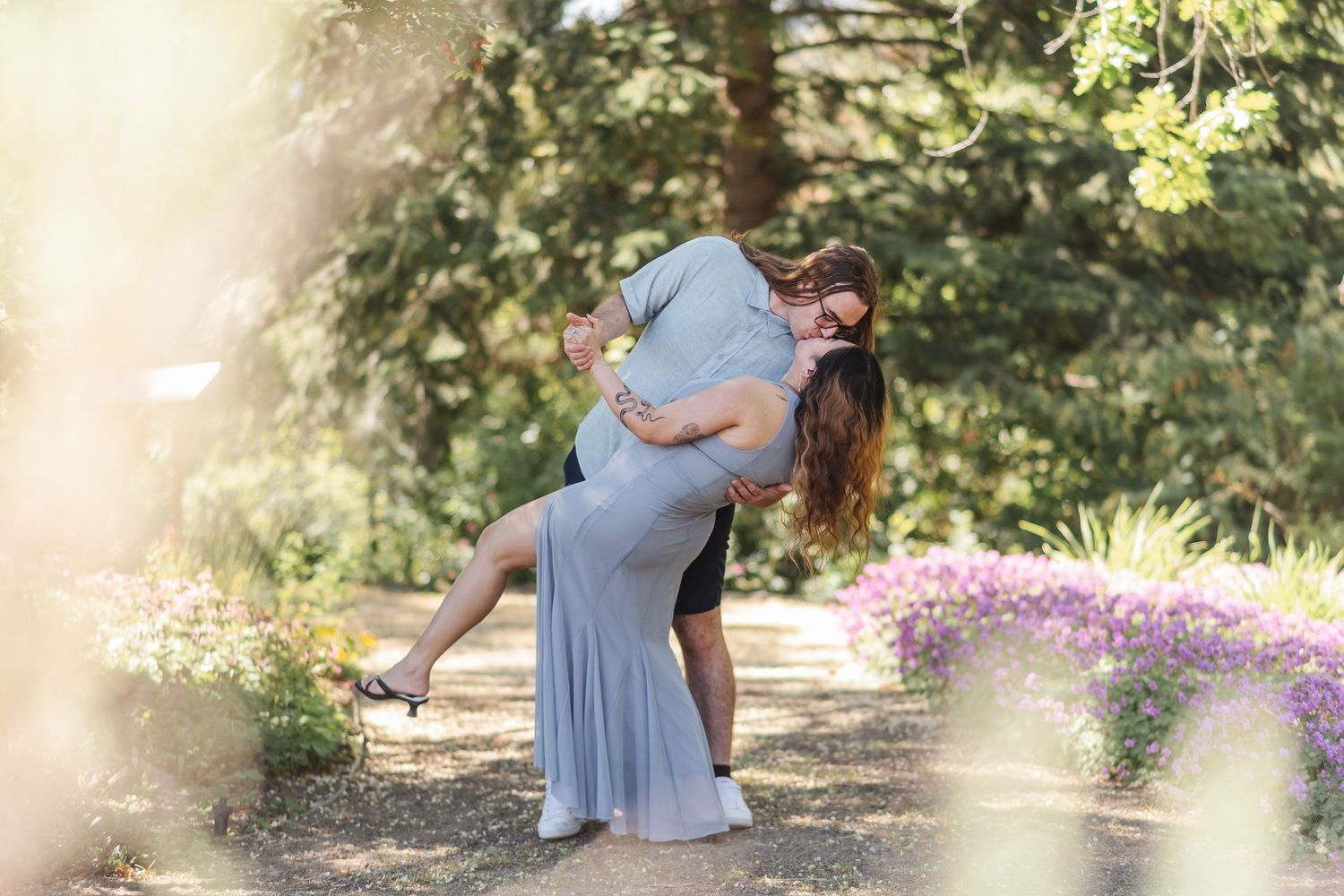 A couple embraces in a sunlit garden setting with blooming flowers and trees creating a romantic outdoor atmosphere.
