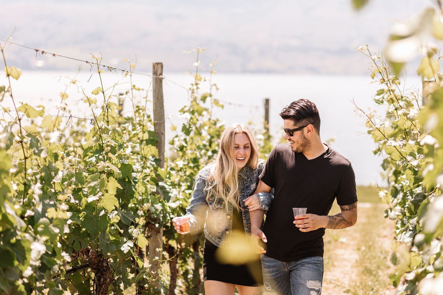 Couple enjoying wine and laughing together while standing in a sunny vineyard with grapevines.