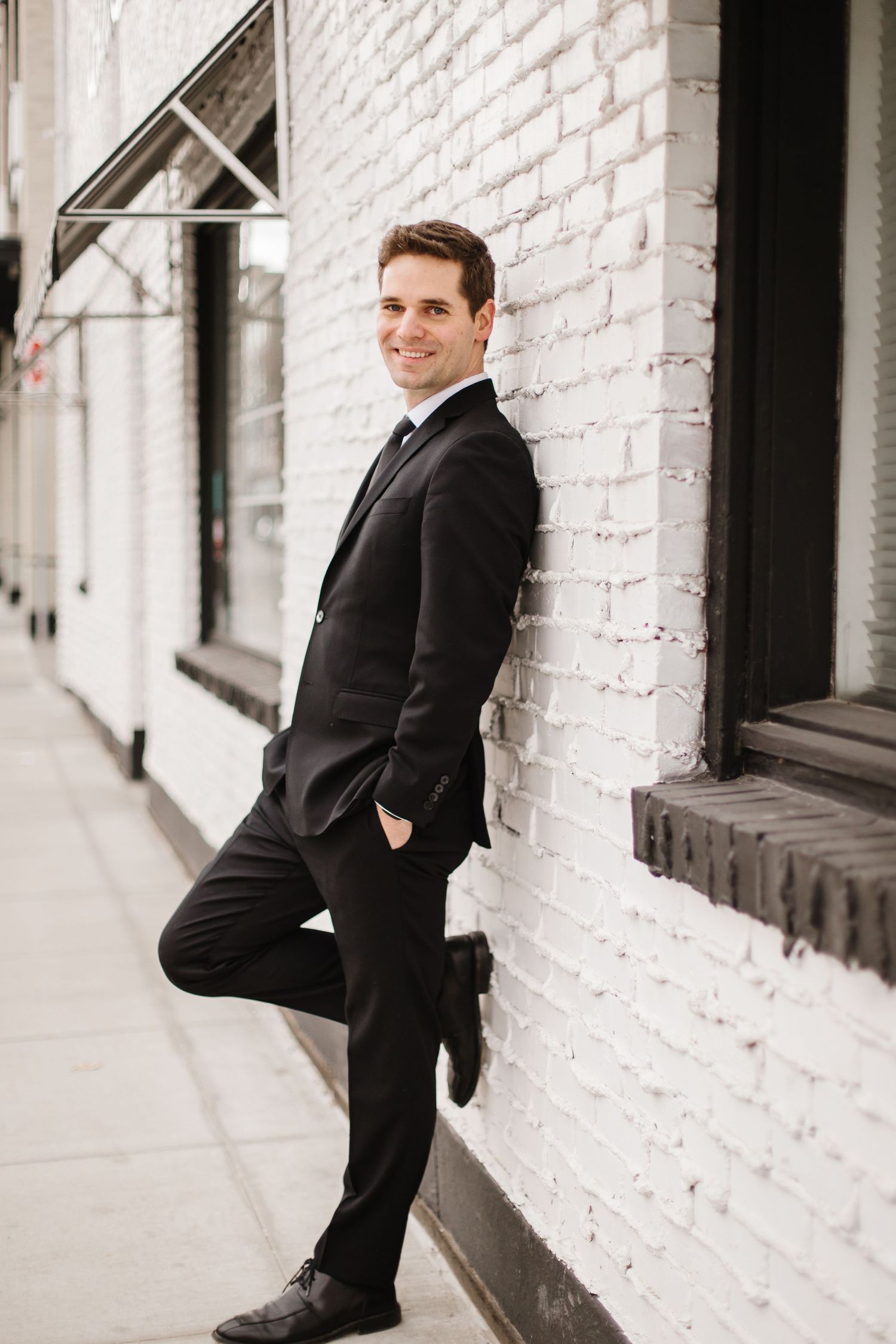 A person in a black suit poses against a white brick wall in a series of professional portrait shots.
