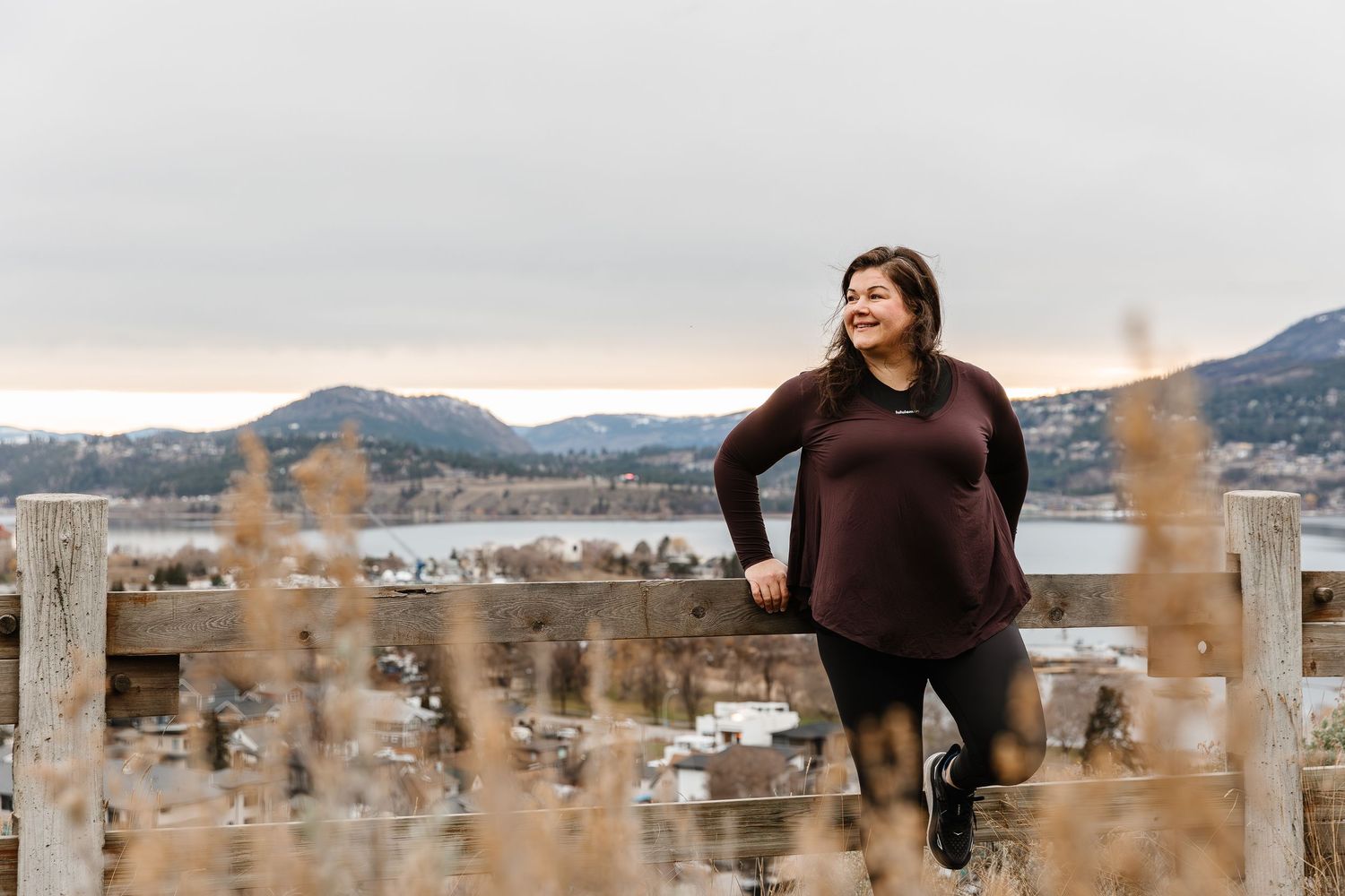 A person in a brown sweater poses in front of snow-capped mountains during a winter sunset.