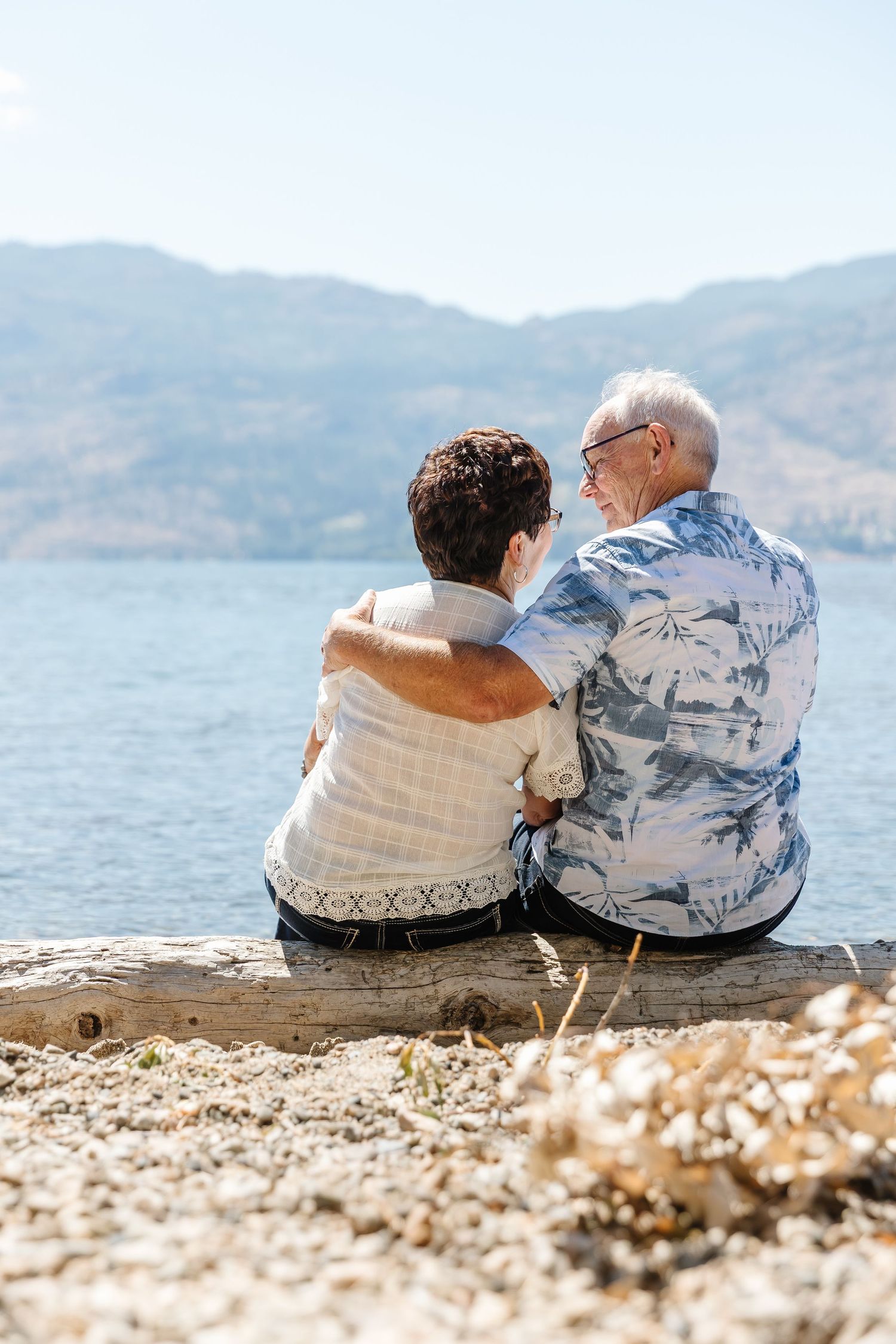 Elderly couple sitting together on rocky beach shore overlooking mountain lake on sunny day.