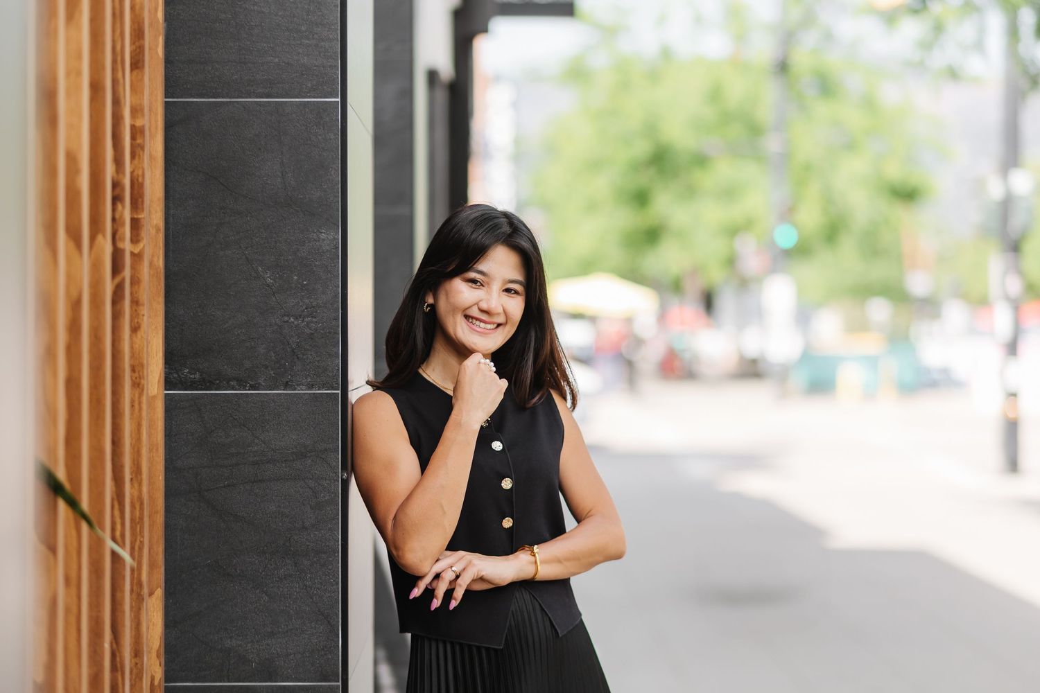 A woman in a black sleeveless dress poses against a wooden doorframe in an outdoor setting.