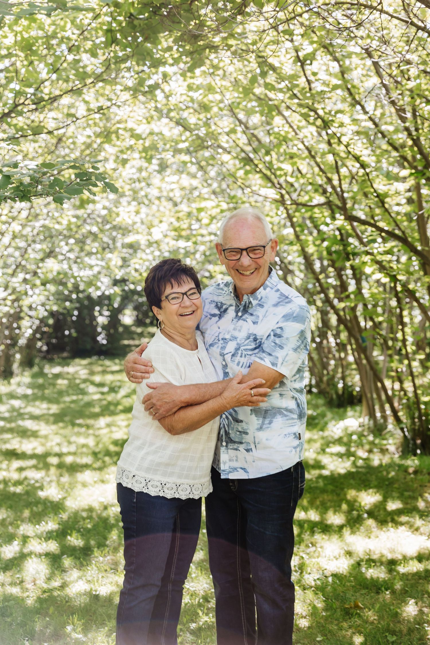 An elderly couple shares a tender embrace while dancing together in a sunlit garden setting with blooming trees.