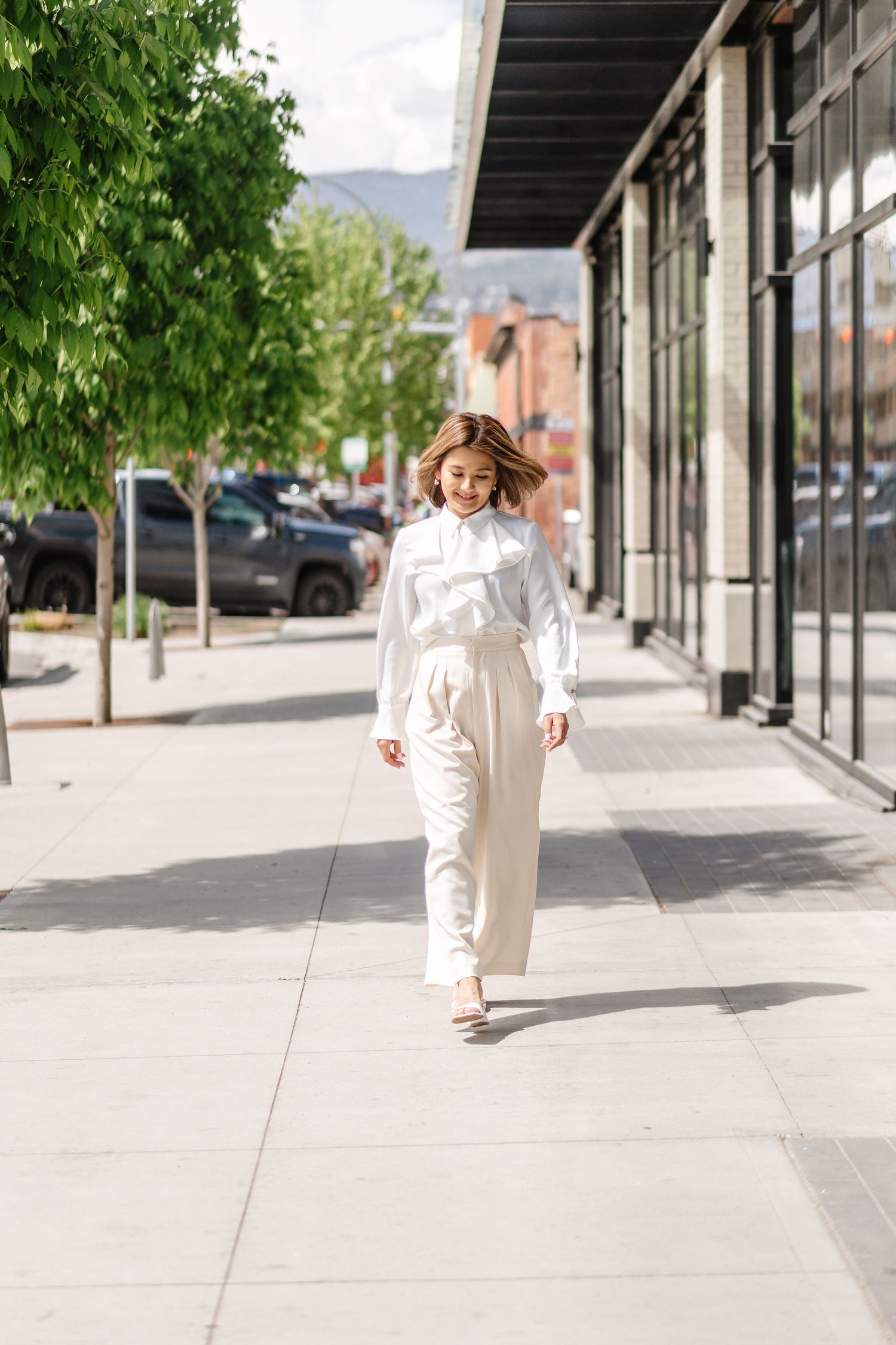 A person in an all-white outfit walks along a modern urban sidewalk with storefronts and trees in the background.