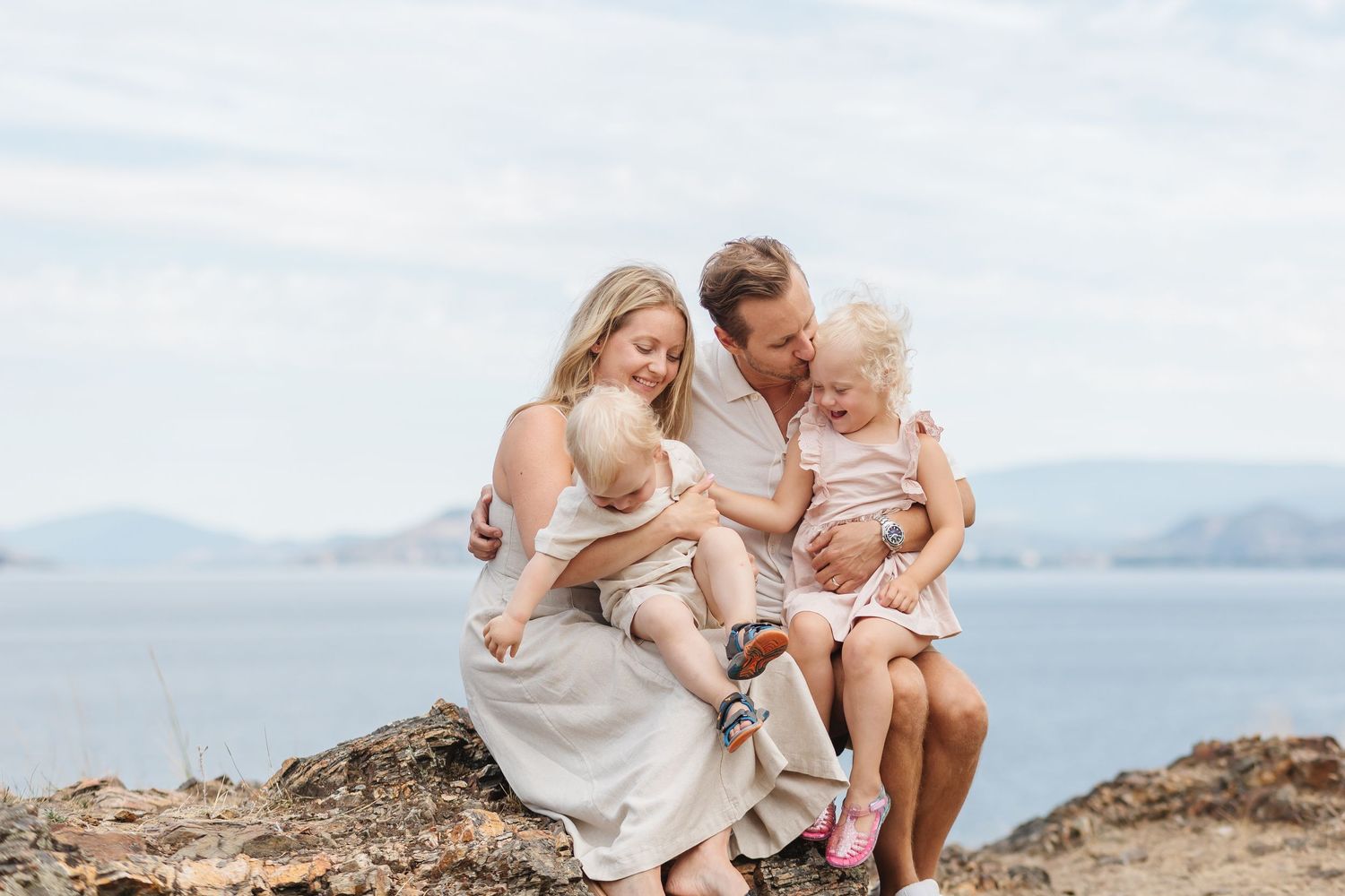 A family sits together on a rocky cliff overlooking a serene lake with mountains in the background.