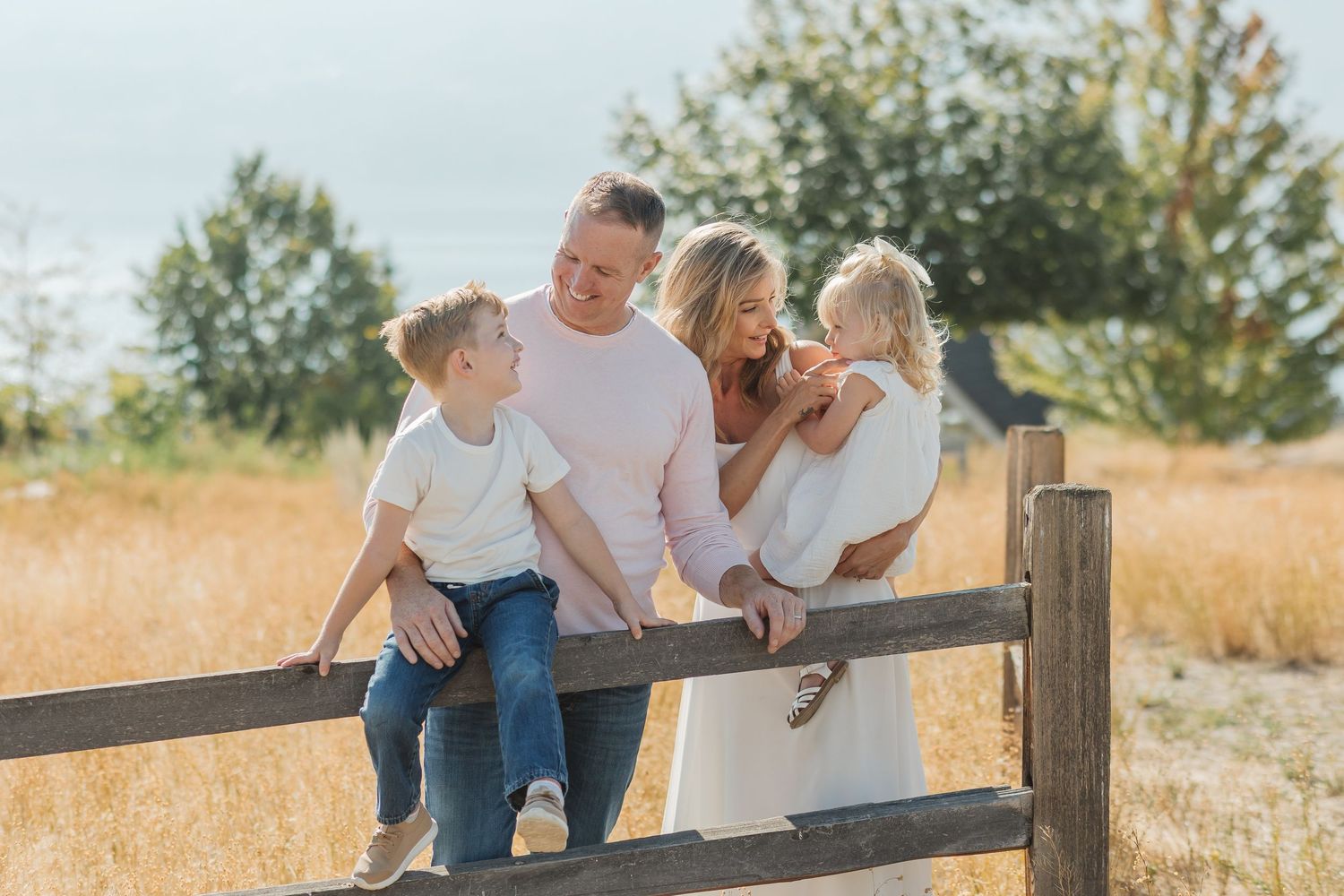 Family in casual white outfits leaning on wooden fence in golden field during sunset photo session.