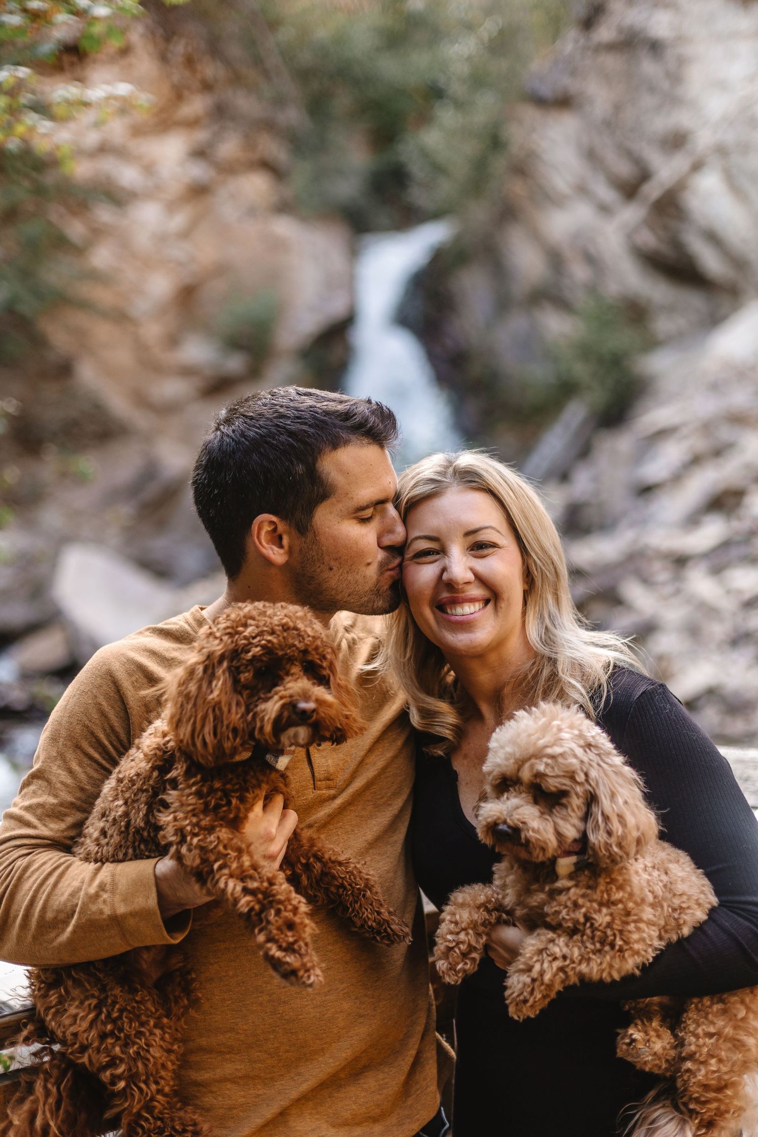 A couple shares tender moments with their brown poodle dog in a scenic outdoor setting with rocky cliffs.