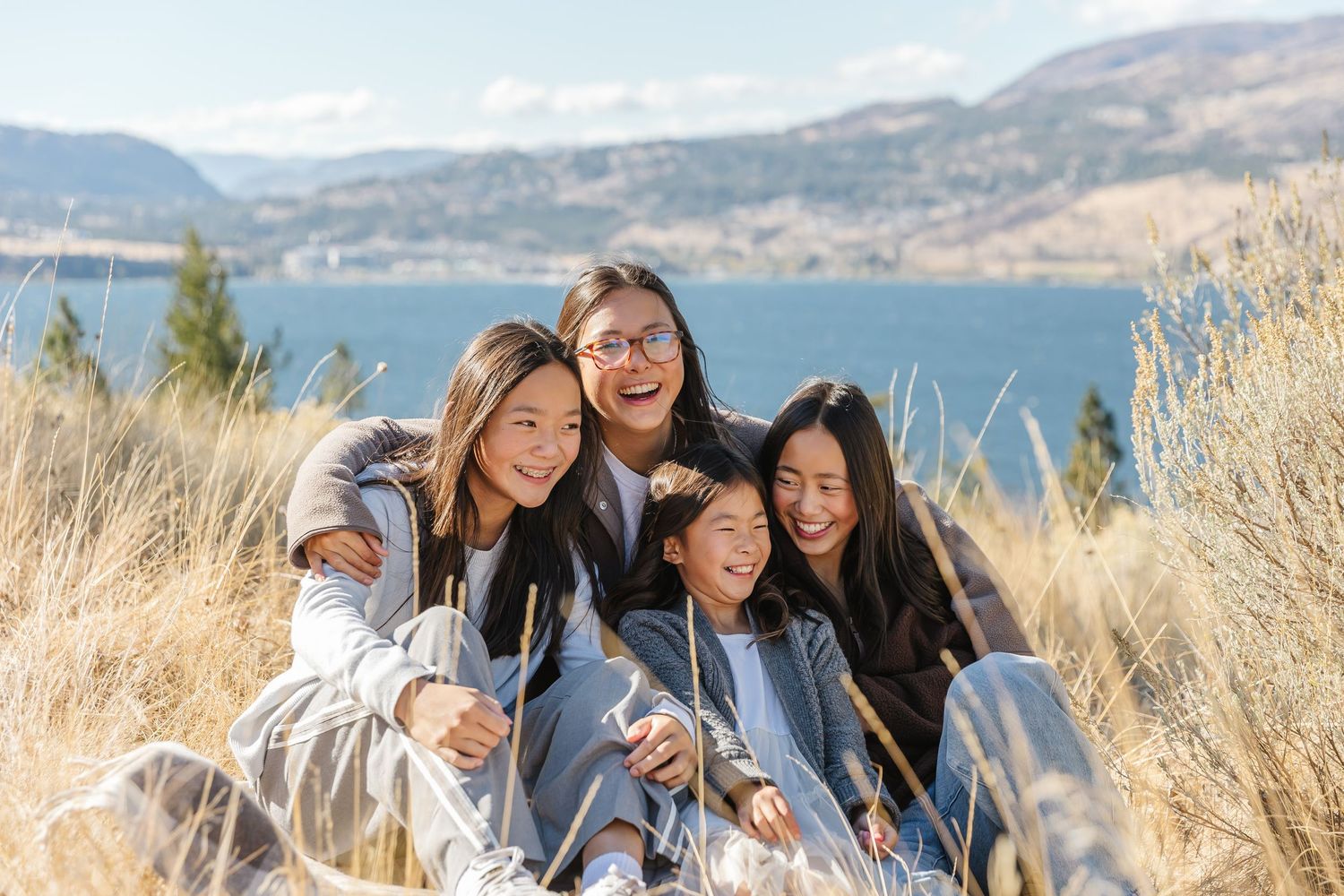 A group of friends laugh together while sitting in tall grass on a hillside overlooking a scenic lake and mountains.