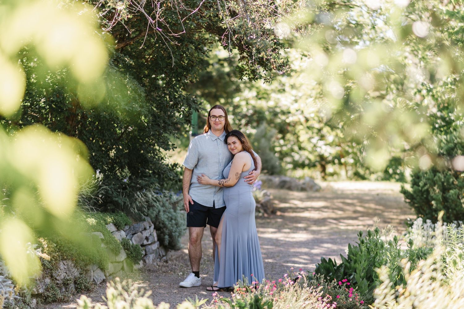 A couple stands together on a sunlit nature trail surrounded by lush greenery and blooming wildflowers.