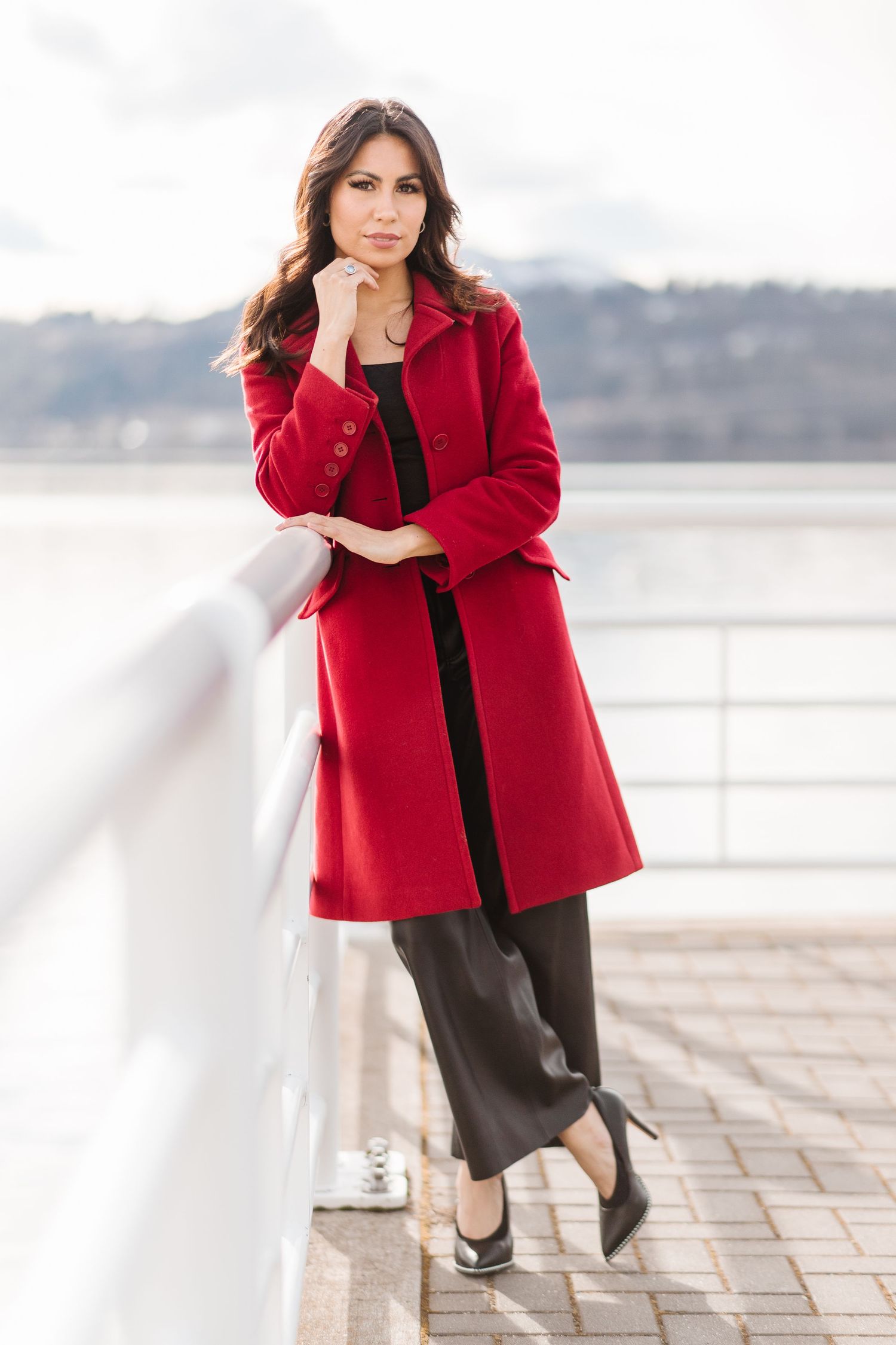 Fashion model in a stylish red coat poses outdoors against a waterfront backdrop.