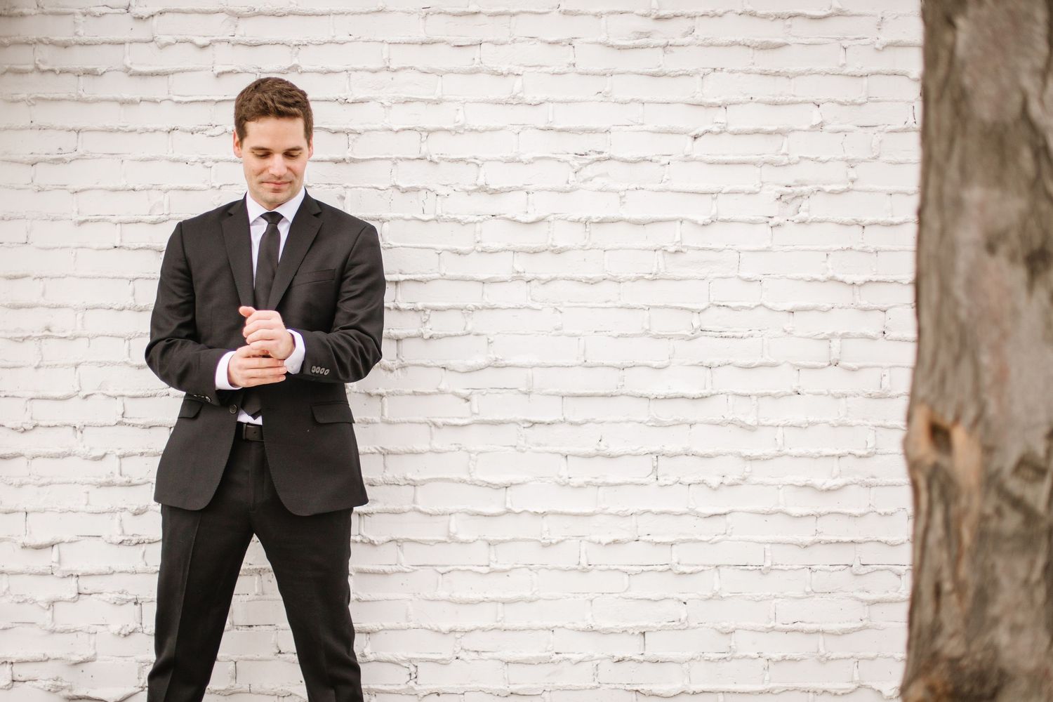A person in a dark suit and tie stands against a white brick wall while looking at their phone.