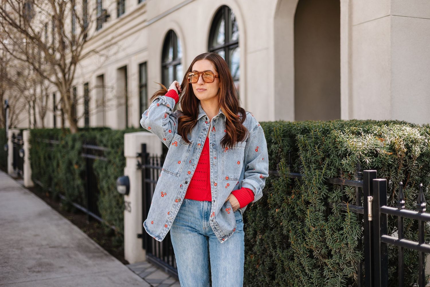 A person wearing a denim jacket and red top poses casually against an urban building exterior with greenery.