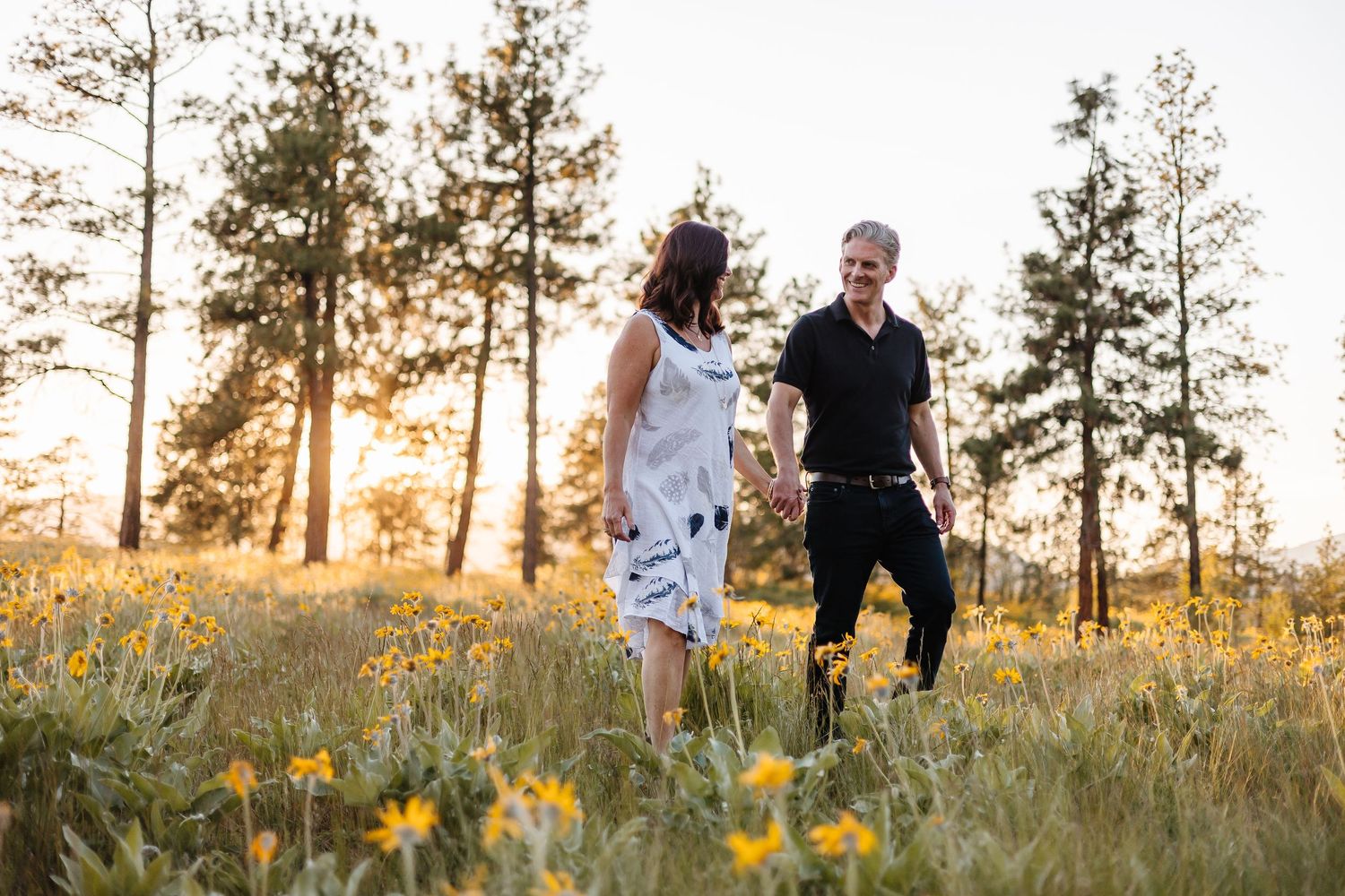A couple walks hand in hand through a golden meadow at sunset with tall pine trees in the background.
