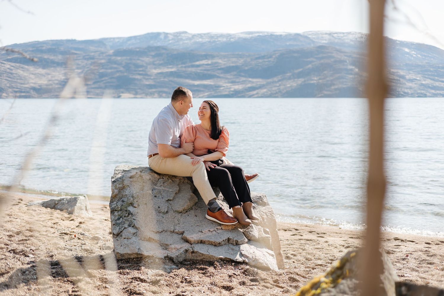 Couple sitting together on a large rock overlooking a serene lake with mountains in the distance.