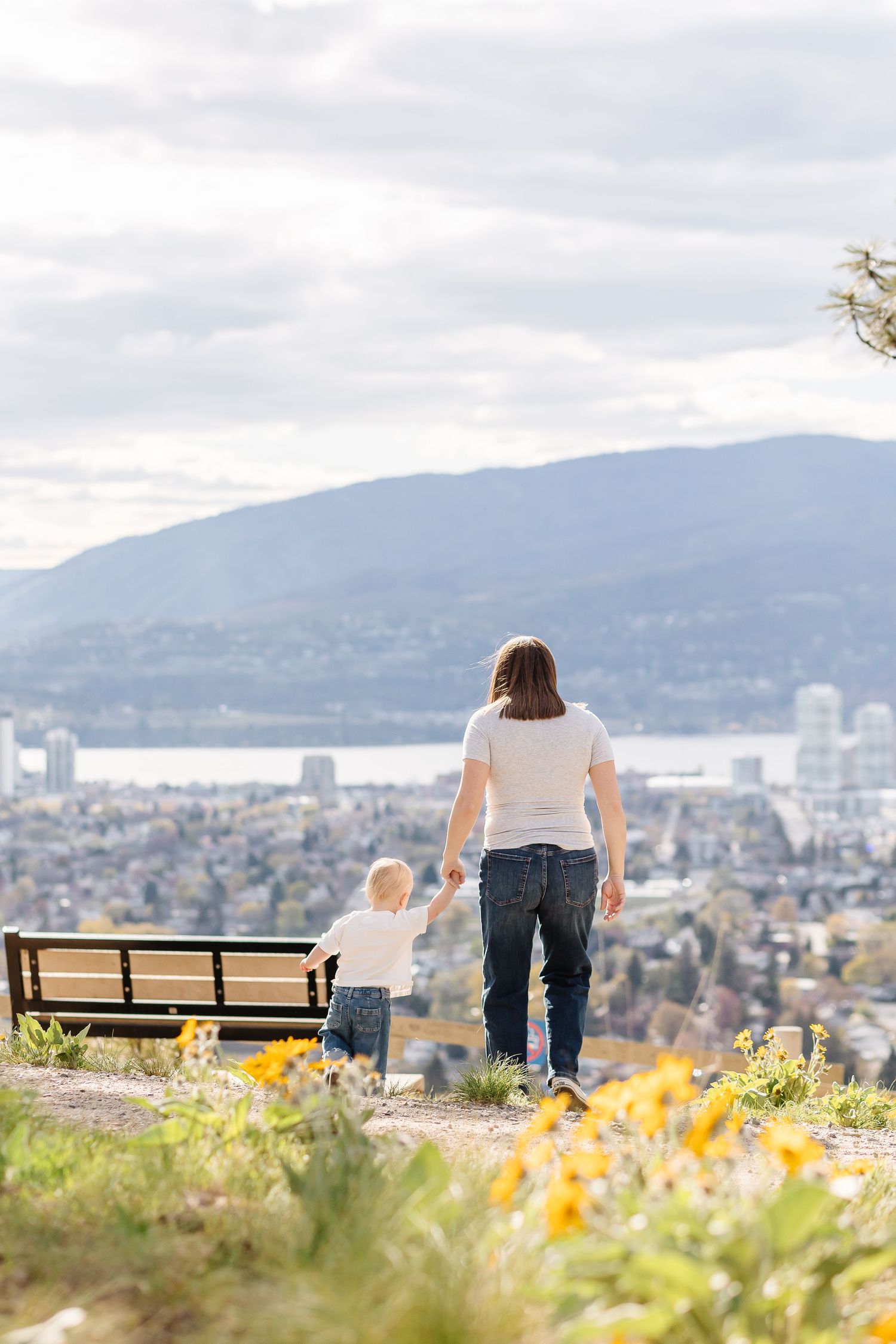 People enjoying a scenic mountain overlook with yellow wildflowers in the foreground and city views below on a cloudy day.