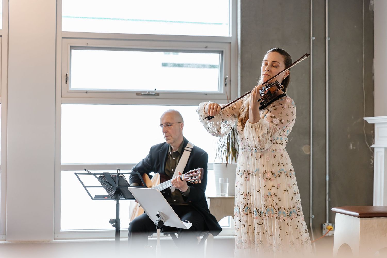 Musicians in formal attire perform together in a bright studio space with large windows.