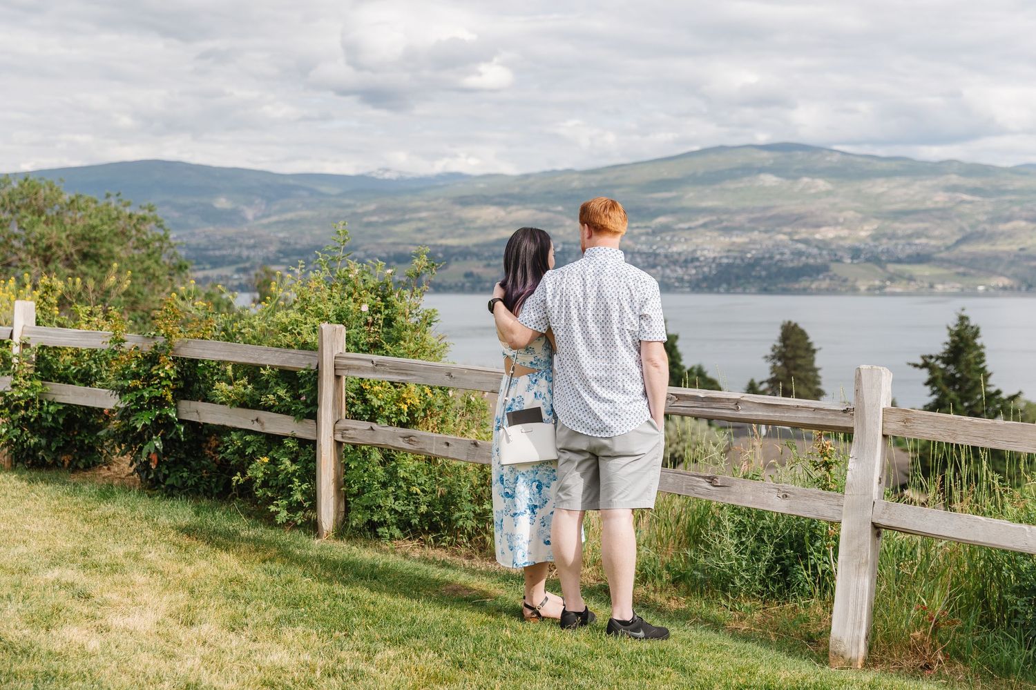 Panoramic view of rolling hills and coastline with wooden fence and visitors enjoying scenic overlook on sunny day.