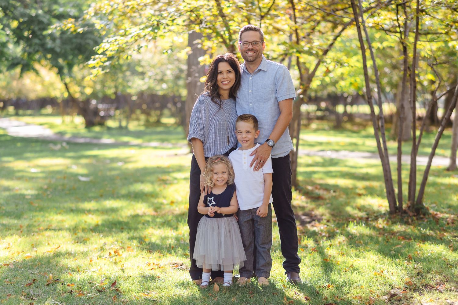 Family poses together in a sunlit autumn park with golden leaves and trees in the background.