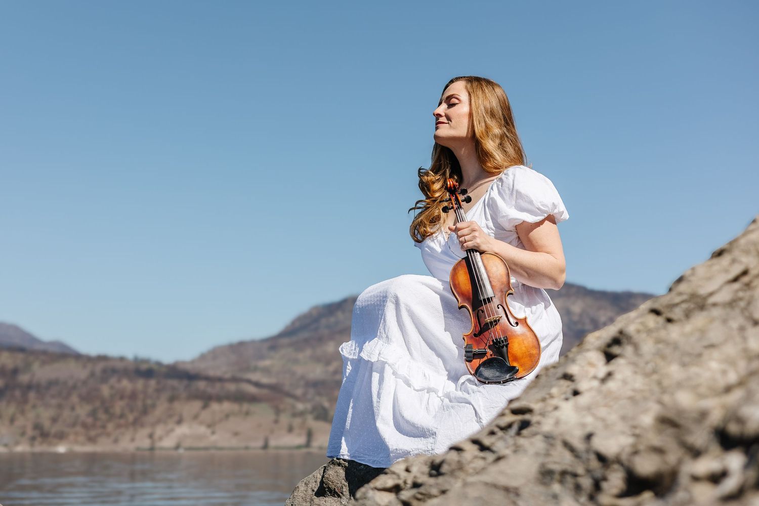 Woman in white dress holding a violin while sitting on rocks near a lake with mountains in the background.