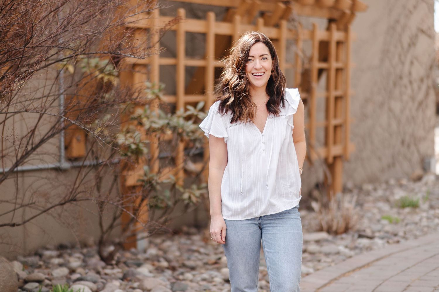 A person in casual attire poses and smiles while standing against a rustic stone wall with wooden lattice decor.