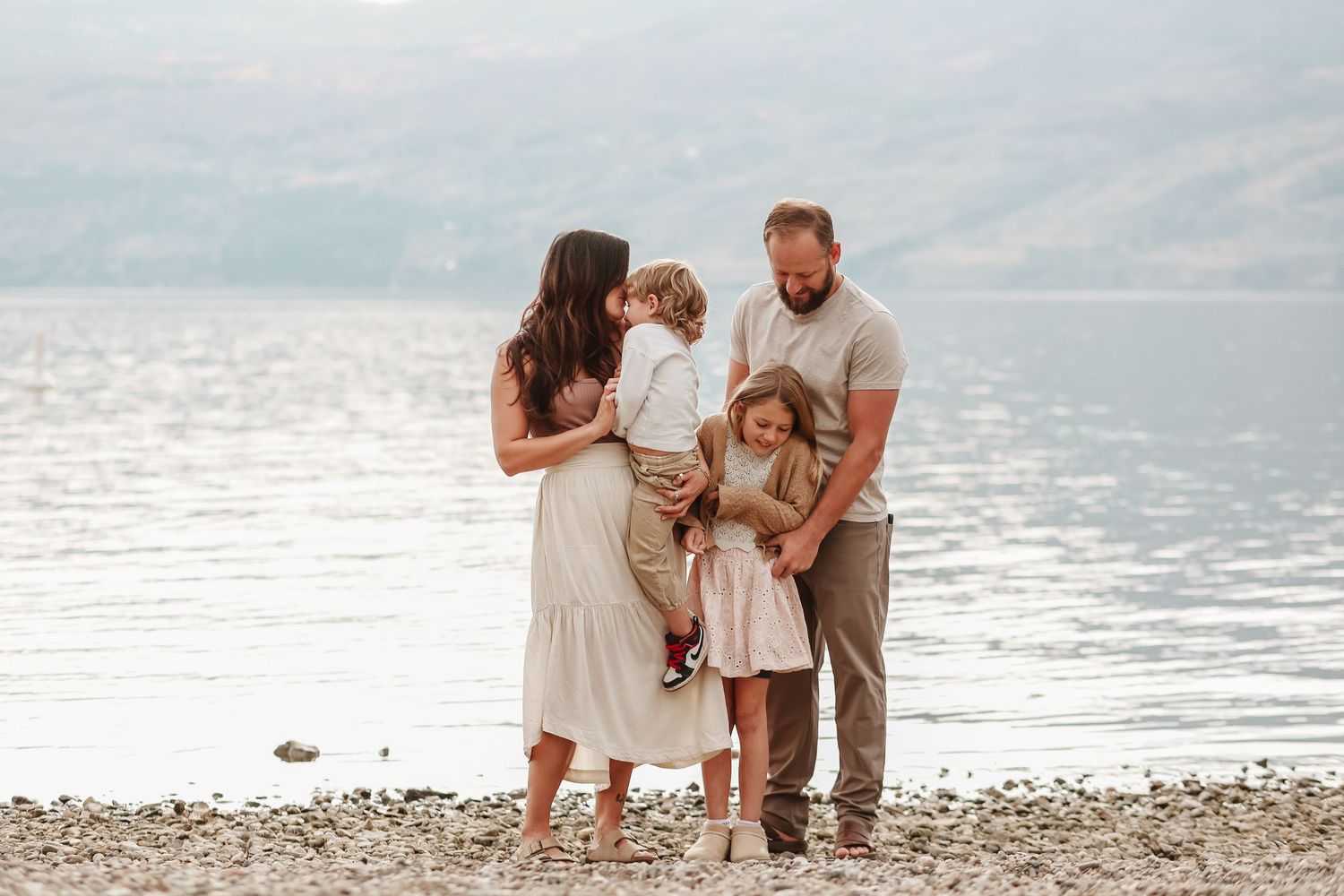 A family in light summer clothing walks together along a rocky beach shoreline at sunset.
