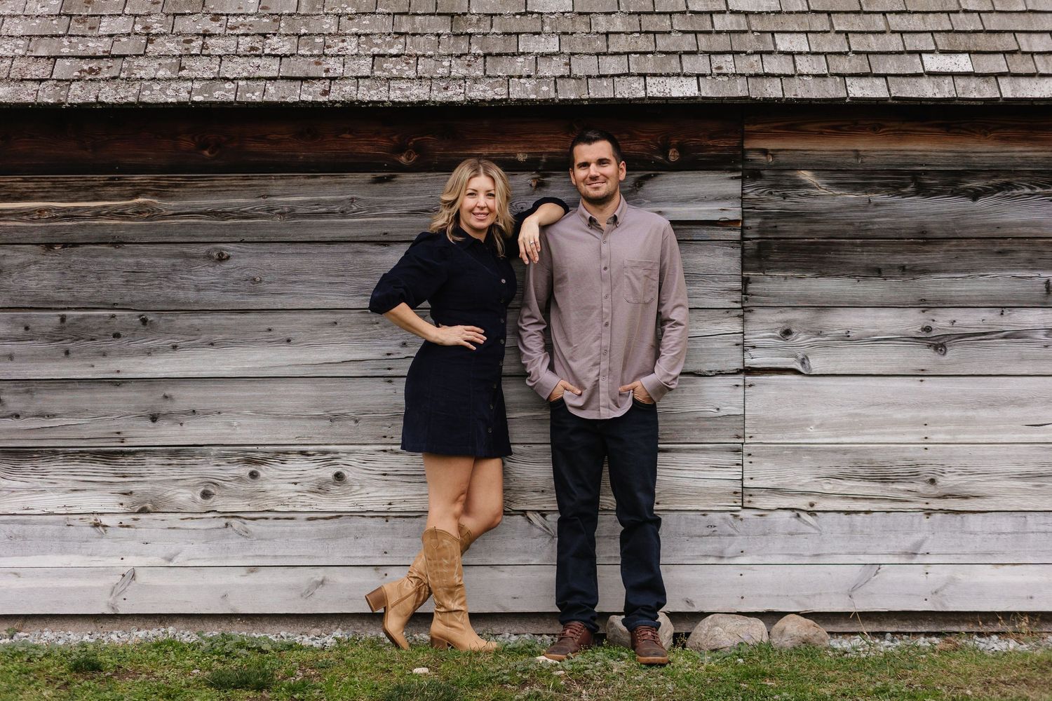 A couple poses in front of a rustic wooden barn wall in casual attire.