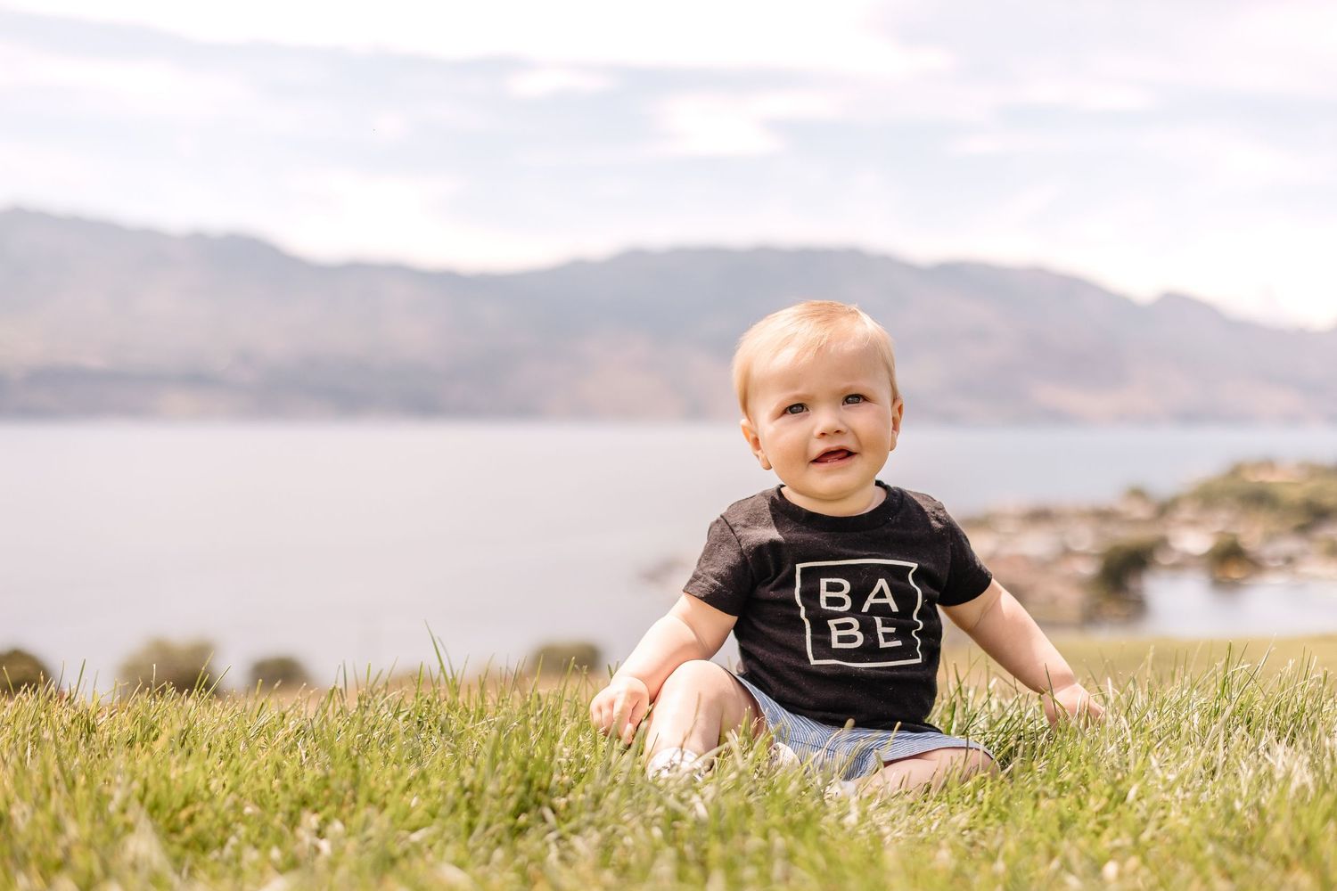 A small child sits in grassy field wearing a black t-shirt with mountains and lake in the background.